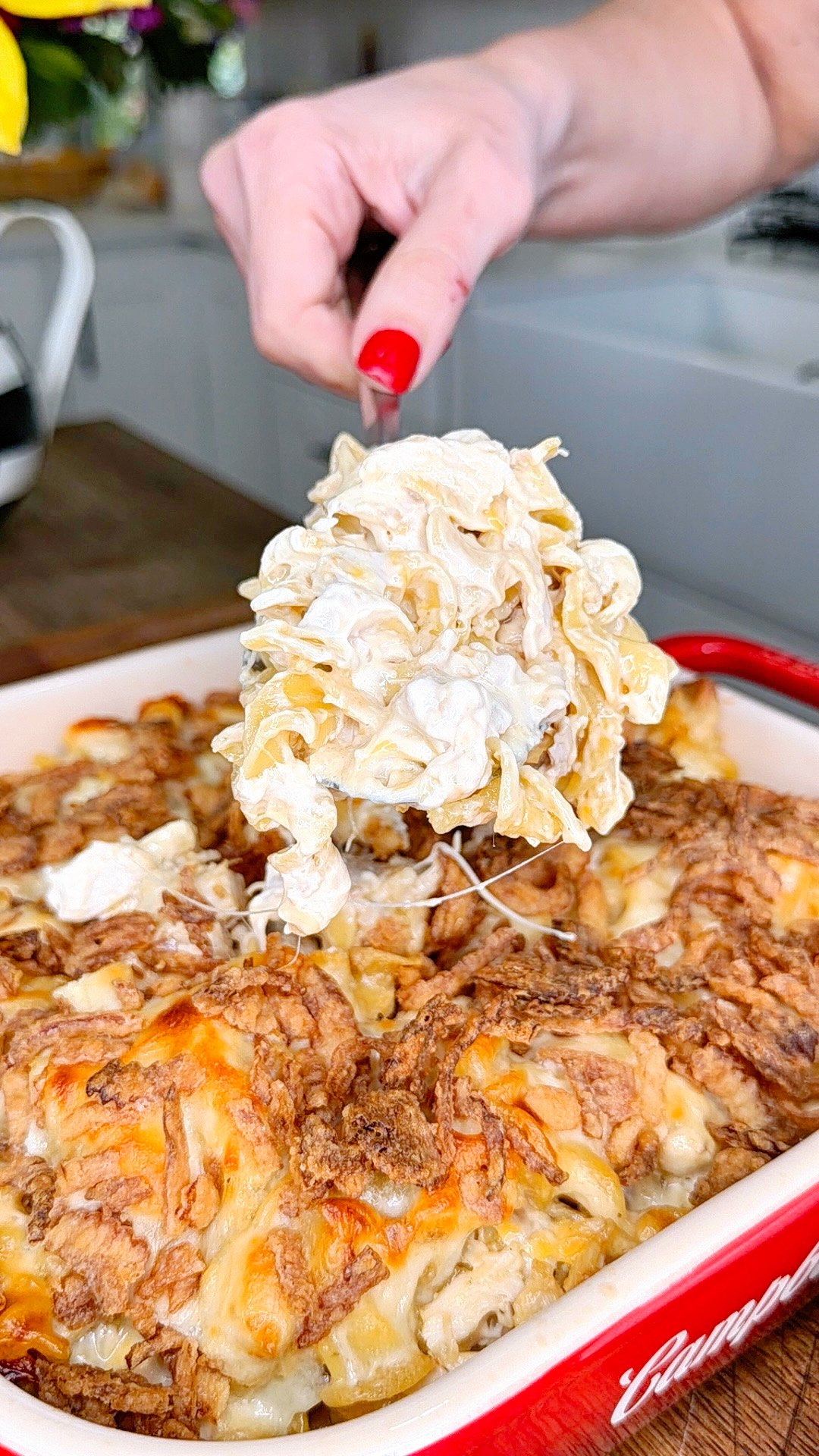 A hand with red nail polish lifts a serving of cheesy, creamy pasta from a baked casserole dish filled with melted, golden-brown cheese. The kitchen background is slightly blurred.