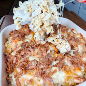 A hand lifts a cheesy, creamy pasta bake from a casserole dish topped with crispy fried onions and melted cheese in a kitchen setting.