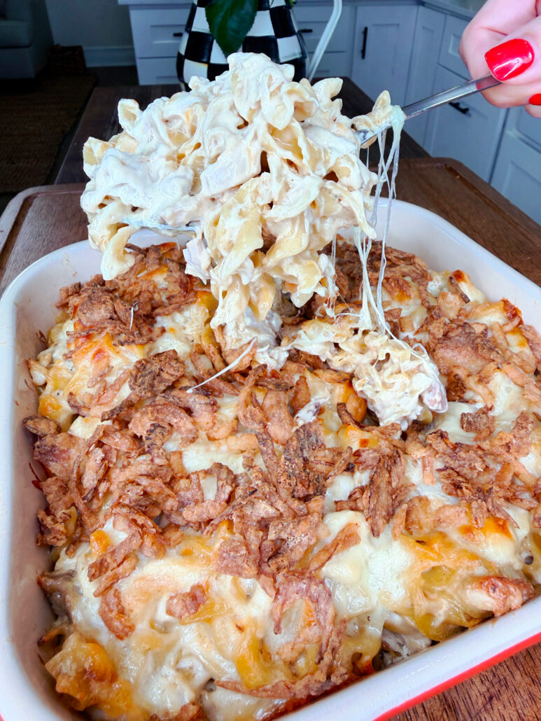 A hand lifts a cheesy, creamy pasta bake from a casserole dish topped with crispy fried onions and melted cheese in a kitchen setting.