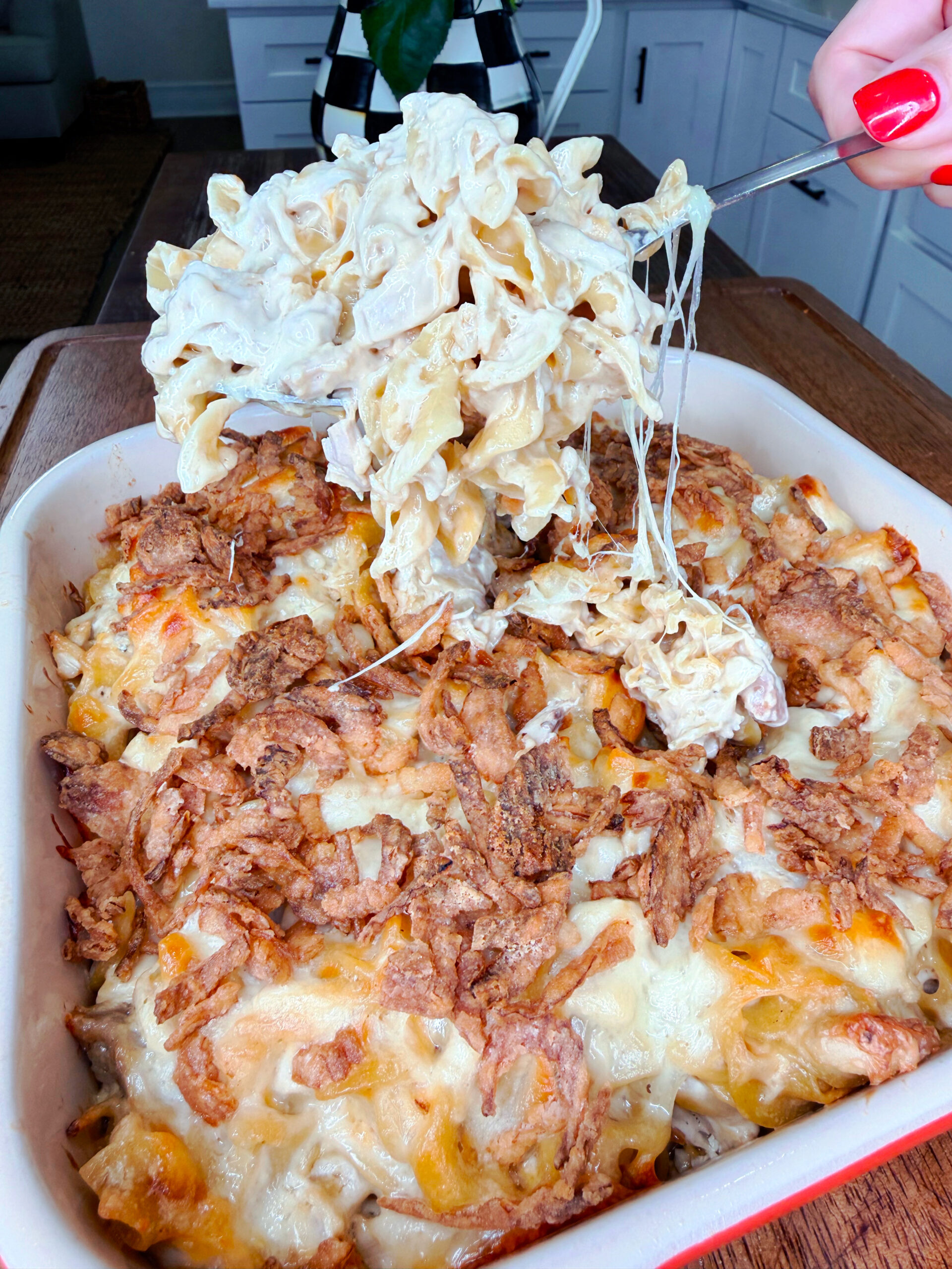 A hand lifts a cheesy, creamy pasta bake from a casserole dish topped with crispy fried onions and melted cheese in a kitchen setting.