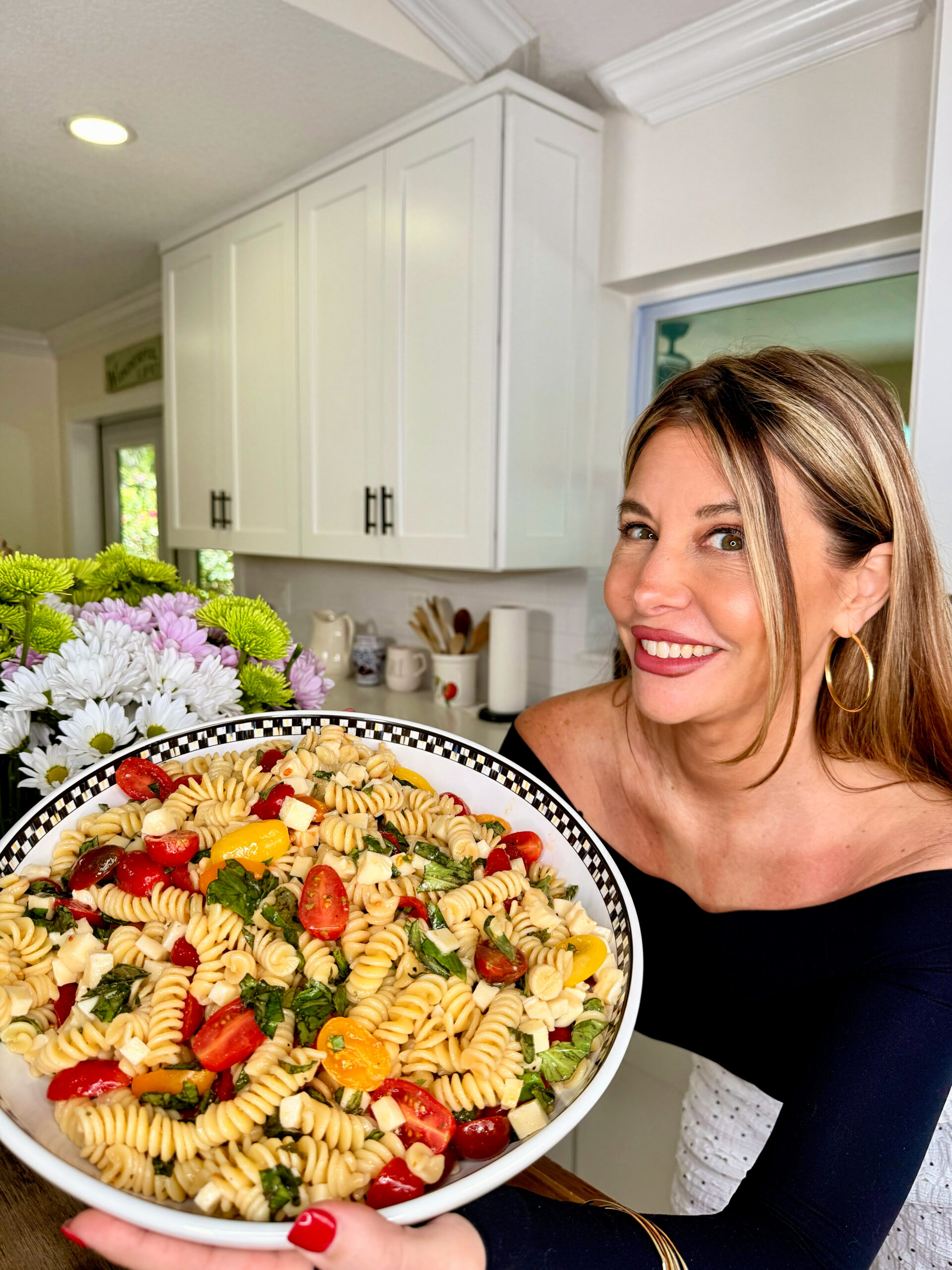 A woman in a black off-the-shoulder top smiles while holding a large bowl of Easy Italian Pasta Salad with tomatoes, basil, and cheese in a bright kitchen with white cabinets and flowers nearby.