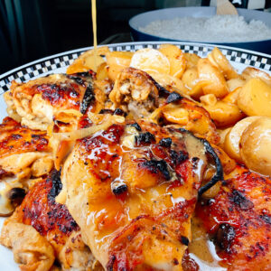 Close-up of honey being drizzled over grilled chicken thighs, reminiscent of One-Pan Mojo Chicken & Potatoes, served with roasted potatoes and pasta on a patterned plate. A bowl of white rice is visible in the background.