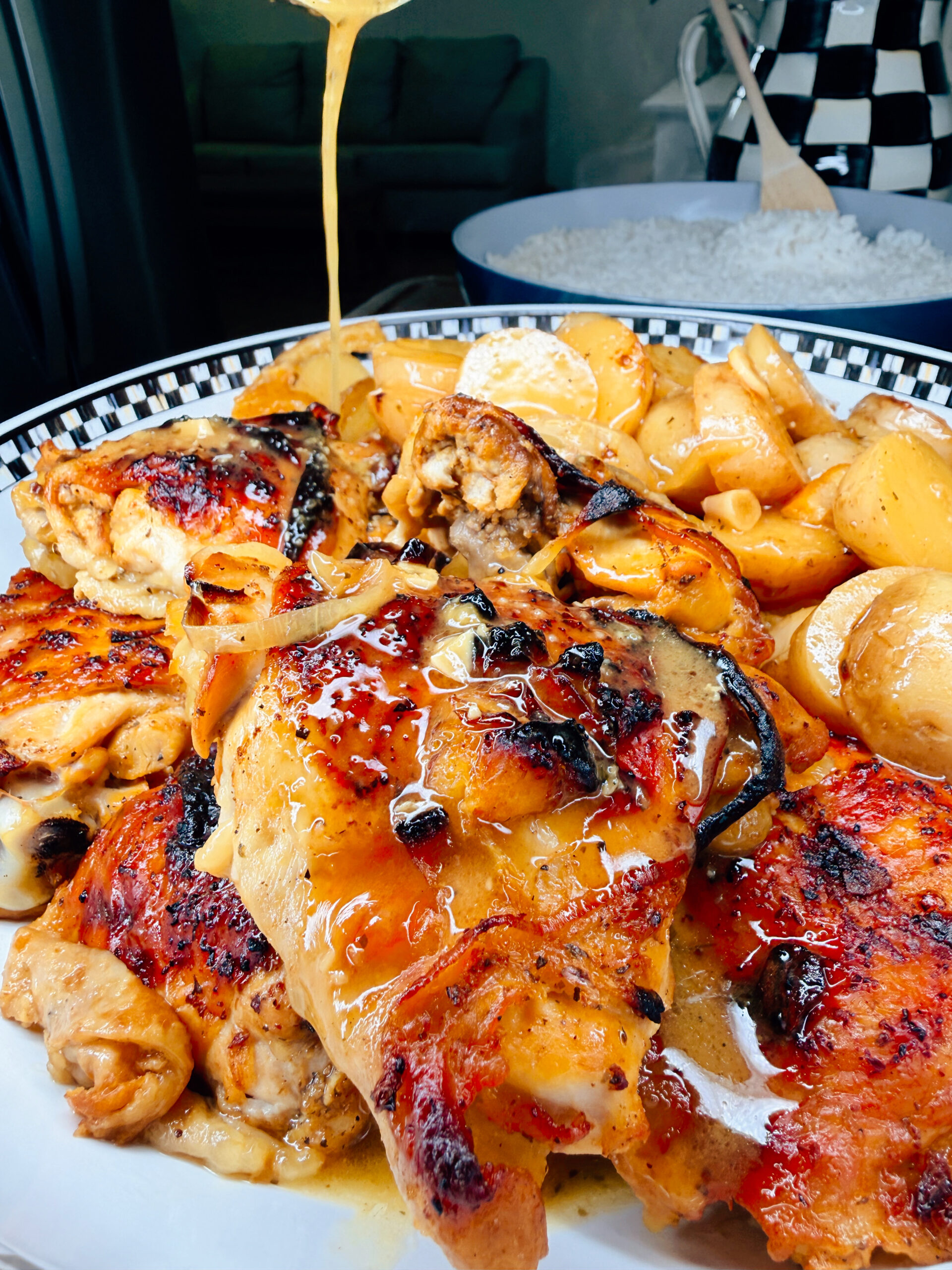 Close-up of honey being drizzled over grilled chicken thighs, reminiscent of One-Pan Mojo Chicken & Potatoes, served with roasted potatoes and pasta on a patterned plate. A bowl of white rice is visible in the background.