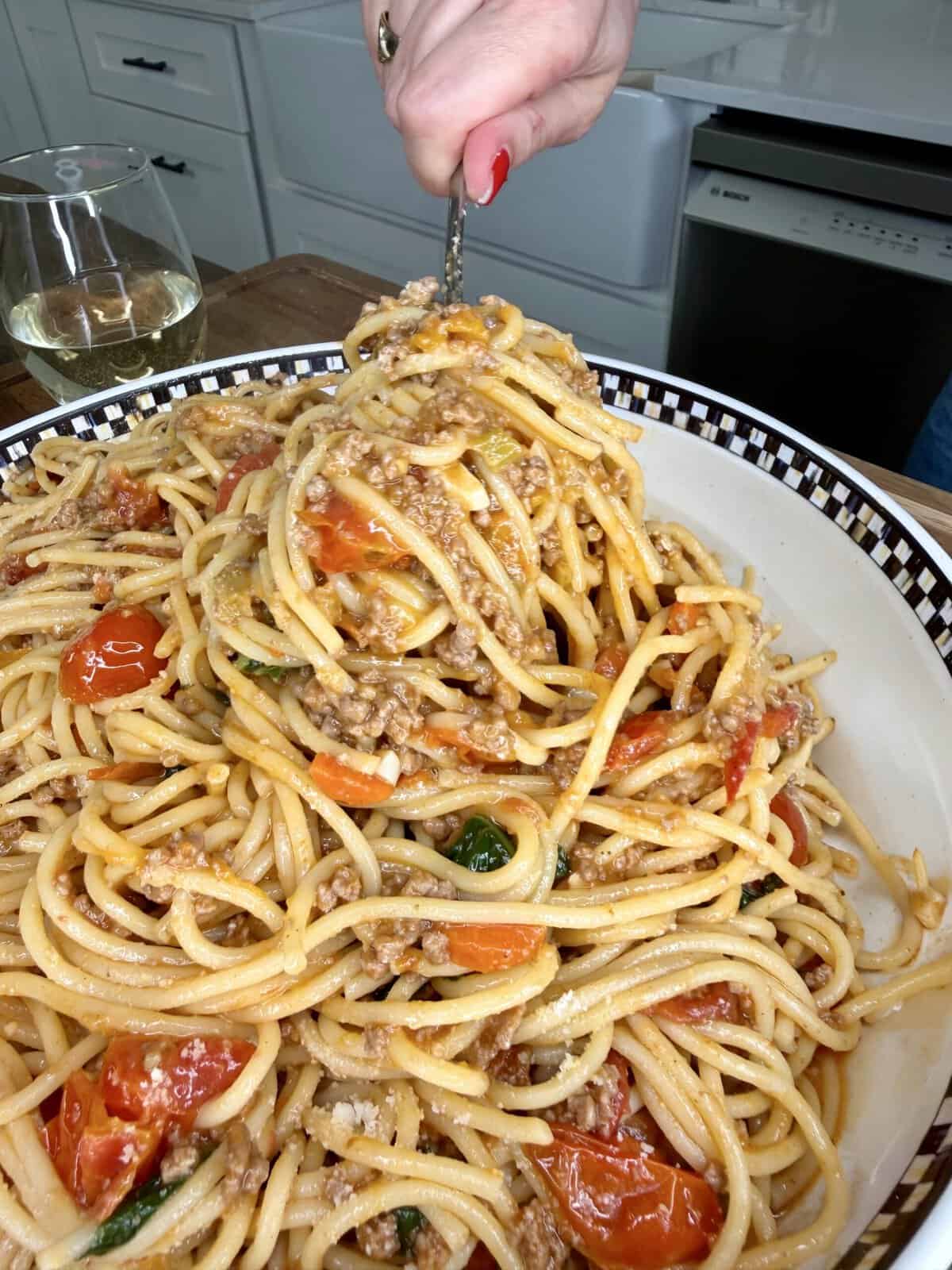 A hand twirls a forkful of spaghetti with meat sauce, cherry tomatoes, and basil in a large bowl. A glass of white wine sits nearby on a wooden countertop in a kitchen setting.