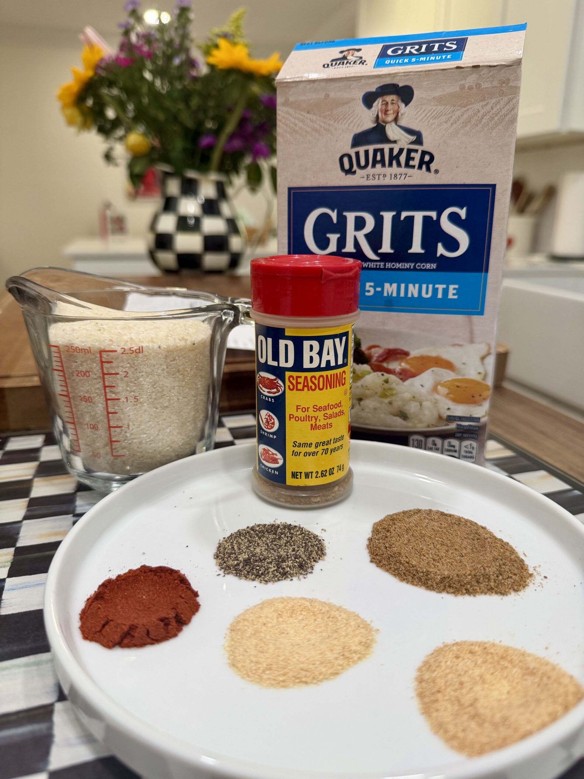 A plate with five spices, a container of Old Bay seasoning, a measuring cup of grits, and a box of Quaker 5-Minute Grits on a kitchen counter, with flowers and a checkered canister in the background.
