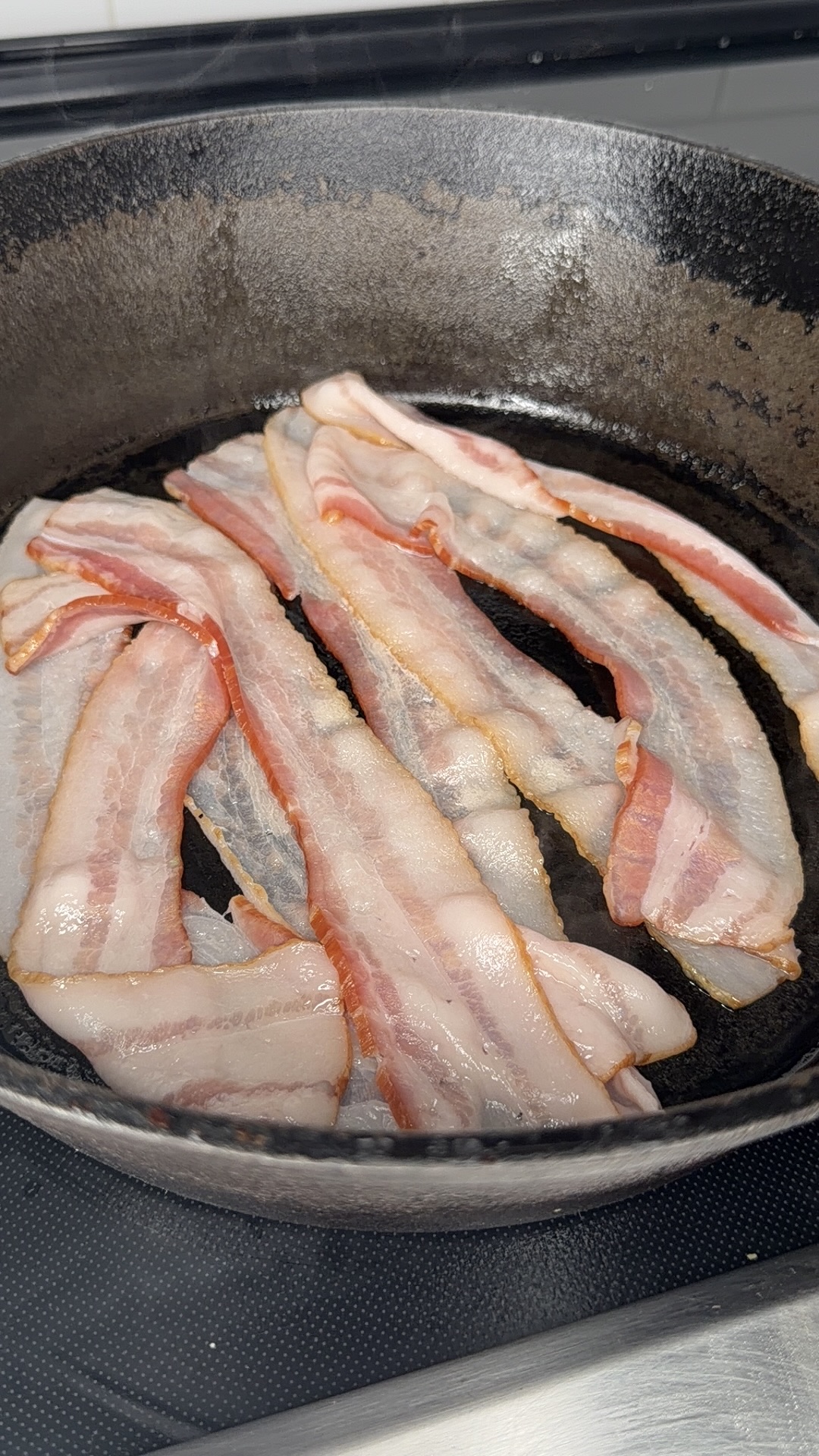 Several raw bacon strips are cooking in a black cast iron skillet on a stovetop.