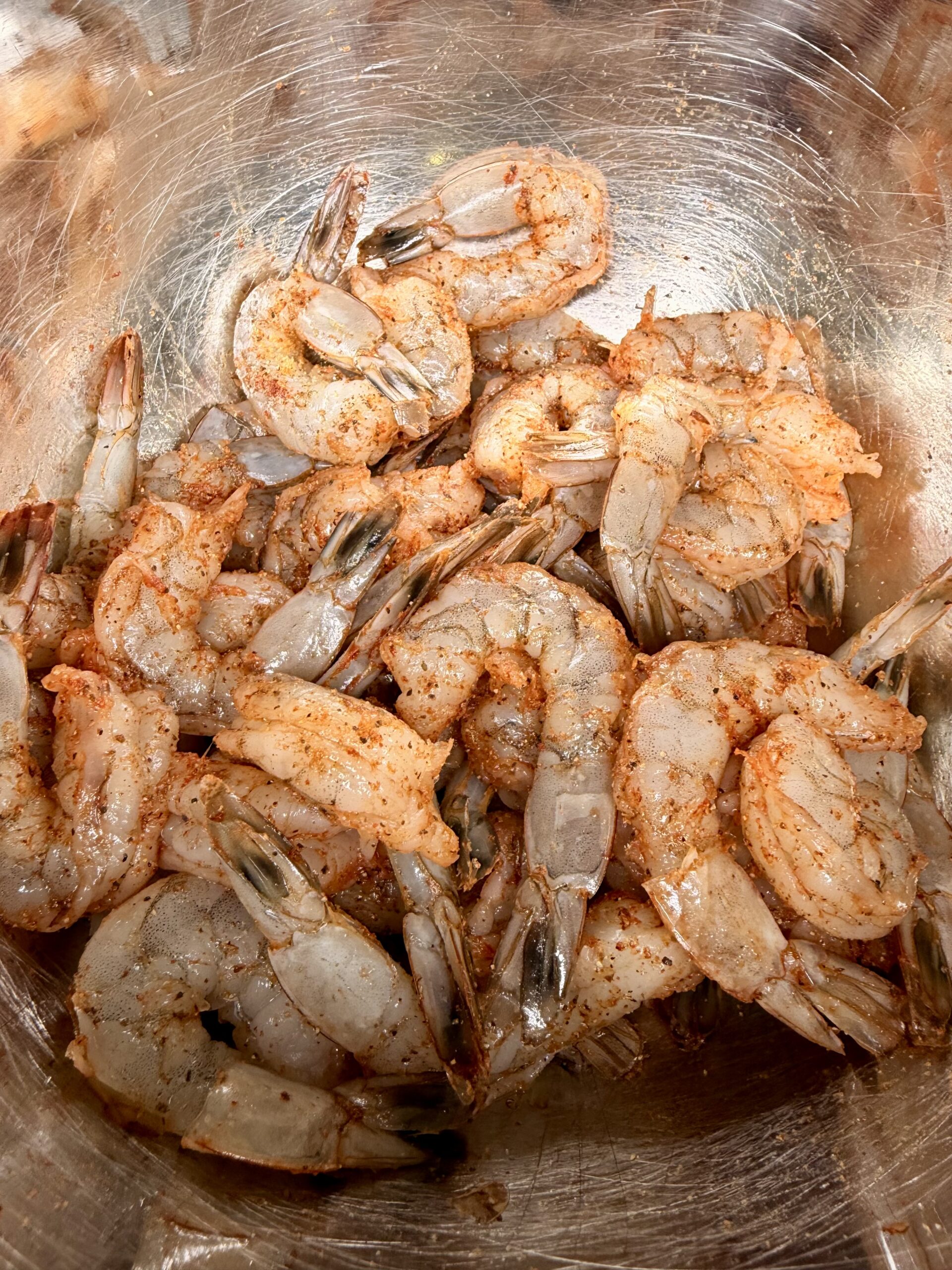 Raw shrimp coated with spices are piled in a metal bowl, ready for cooking. The shrimp are deveined and their shells have been partially removed. The seasoning gives them a reddish tint.