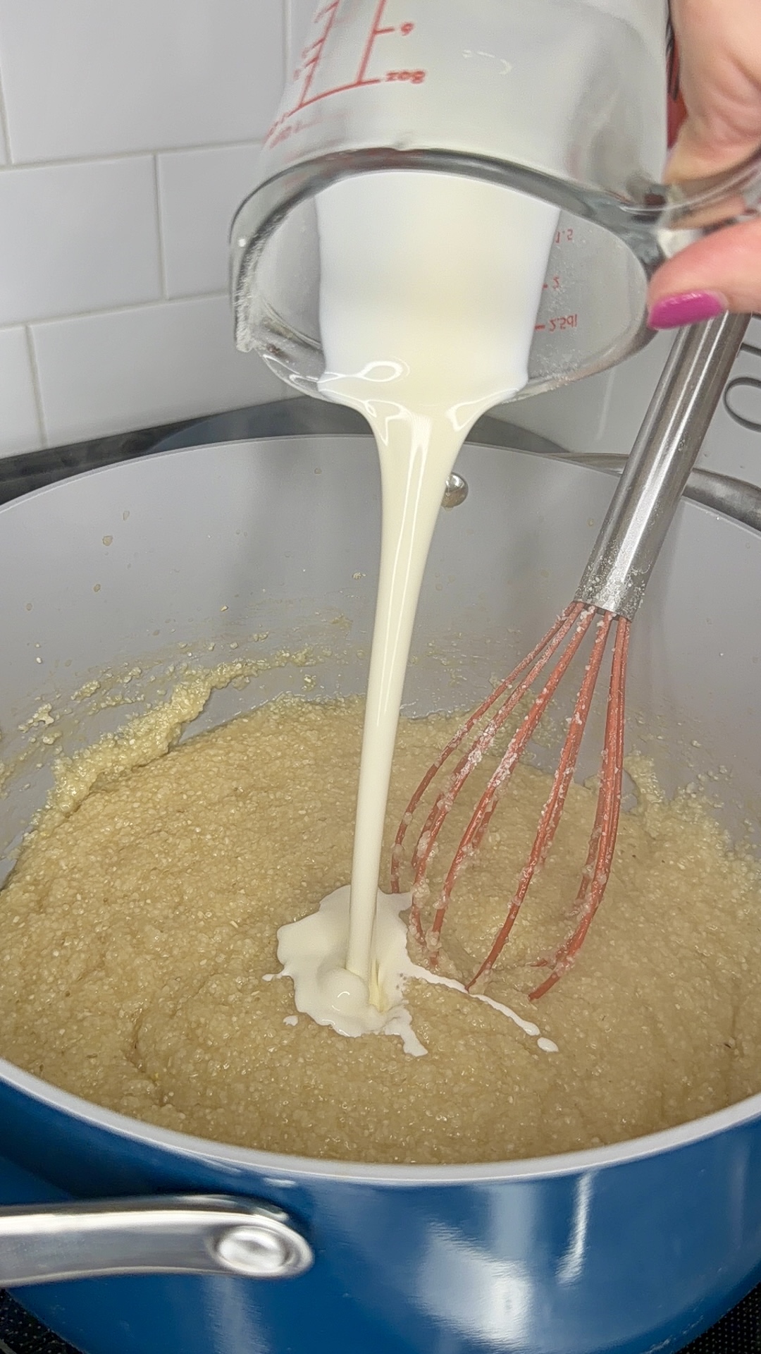 A hand pours milk from a measuring cup into a saucepan of thick, grainy mixture while whisking, on a stovetop with a white tiled backsplash.