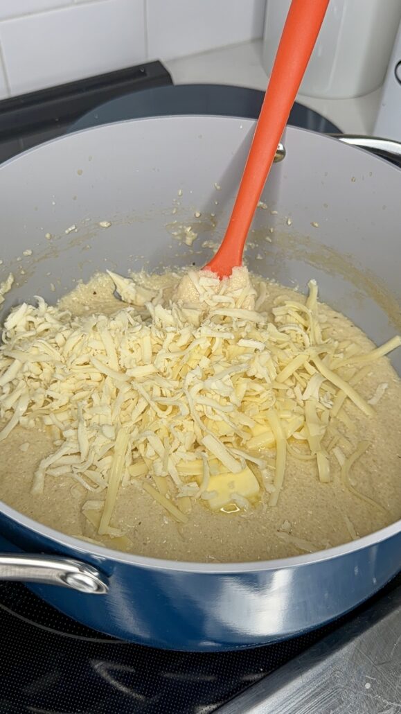 A blue pot on a stove contains Creamy Southern Shrimp & Grits with Bacon Pan Sauce, topped with shredded cheese and butter, being stirred with an orange spatula.