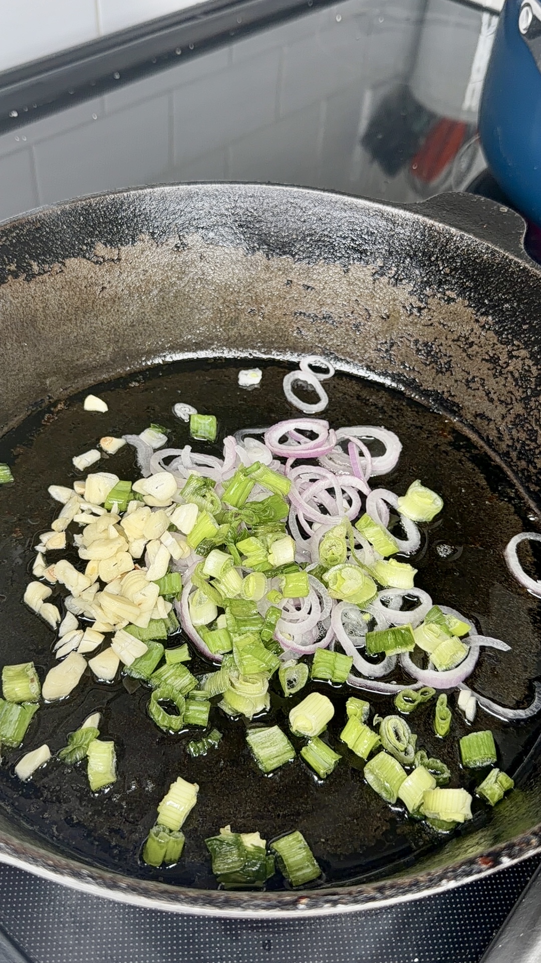 Sliced garlic, green onions, and shallots are being sautéed in a black skillet on a stovetop.