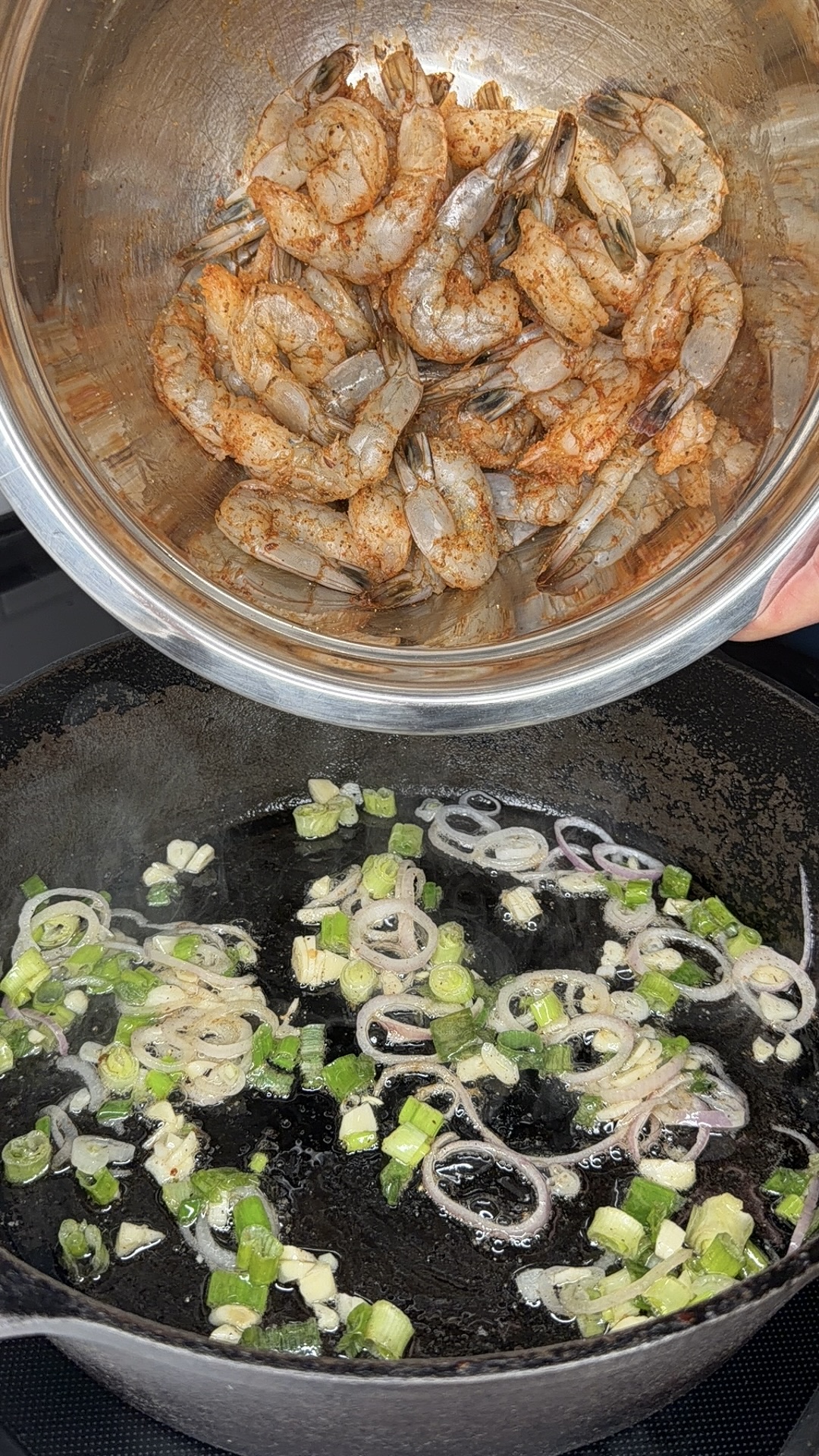 A metal bowl holds raw, seasoned shrimp above a cast iron skillet where sliced shallots, green onions, and garlic are being sautéed in oil. Steam rises from the pan.