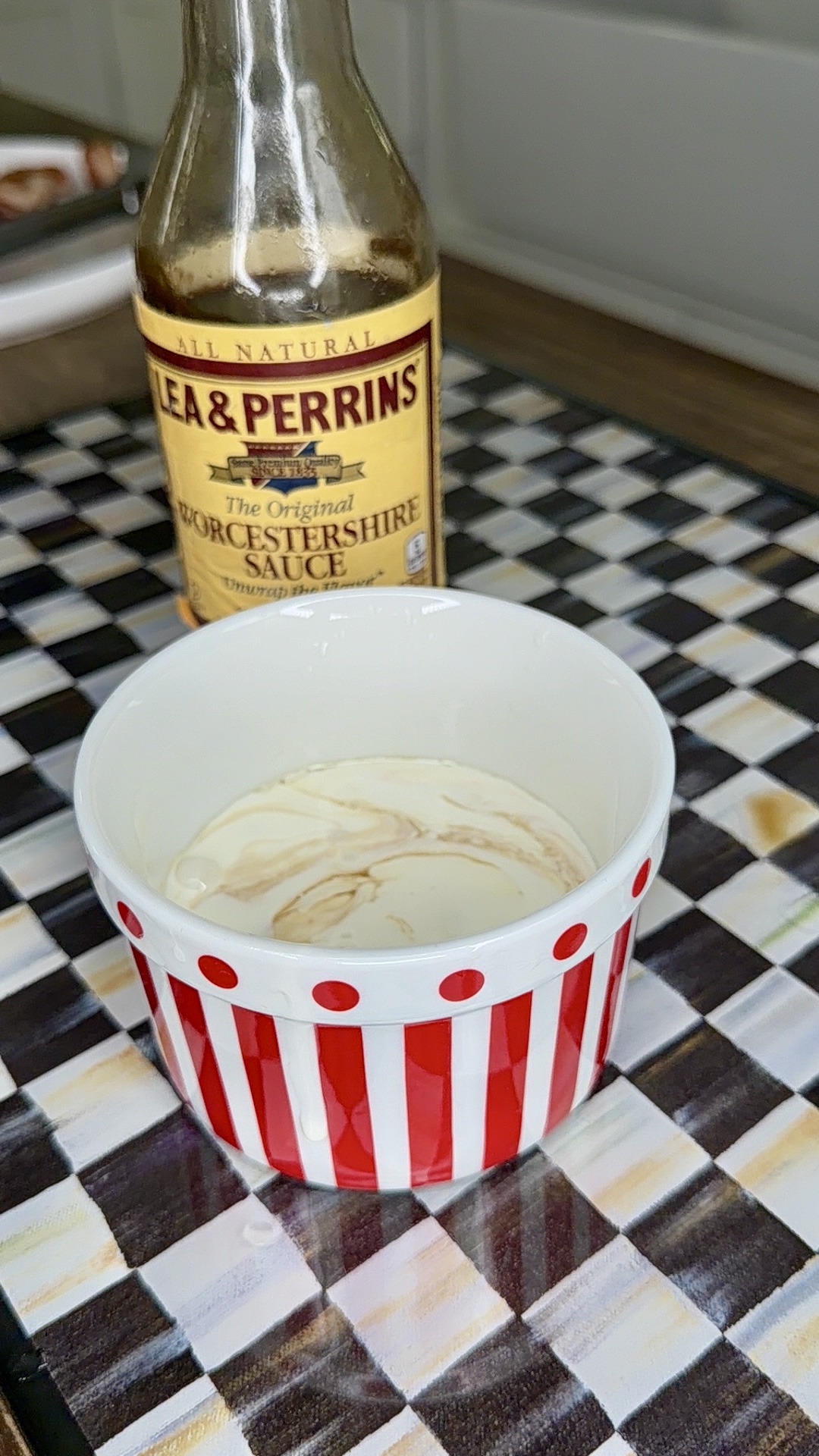 A red and white striped cup with a liquid mixture inside sits on a checkered surface, with a bottle of Lea & Perrins Worcestershire Sauce positioned behind it.