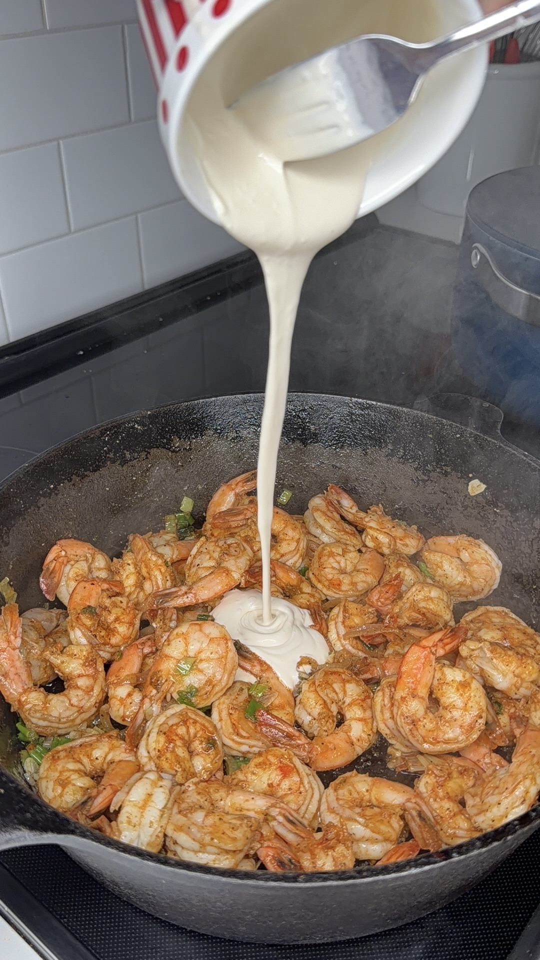A creamy white sauce is being poured from a cup onto seasoned shrimp cooking in a cast iron skillet on a stovetop.