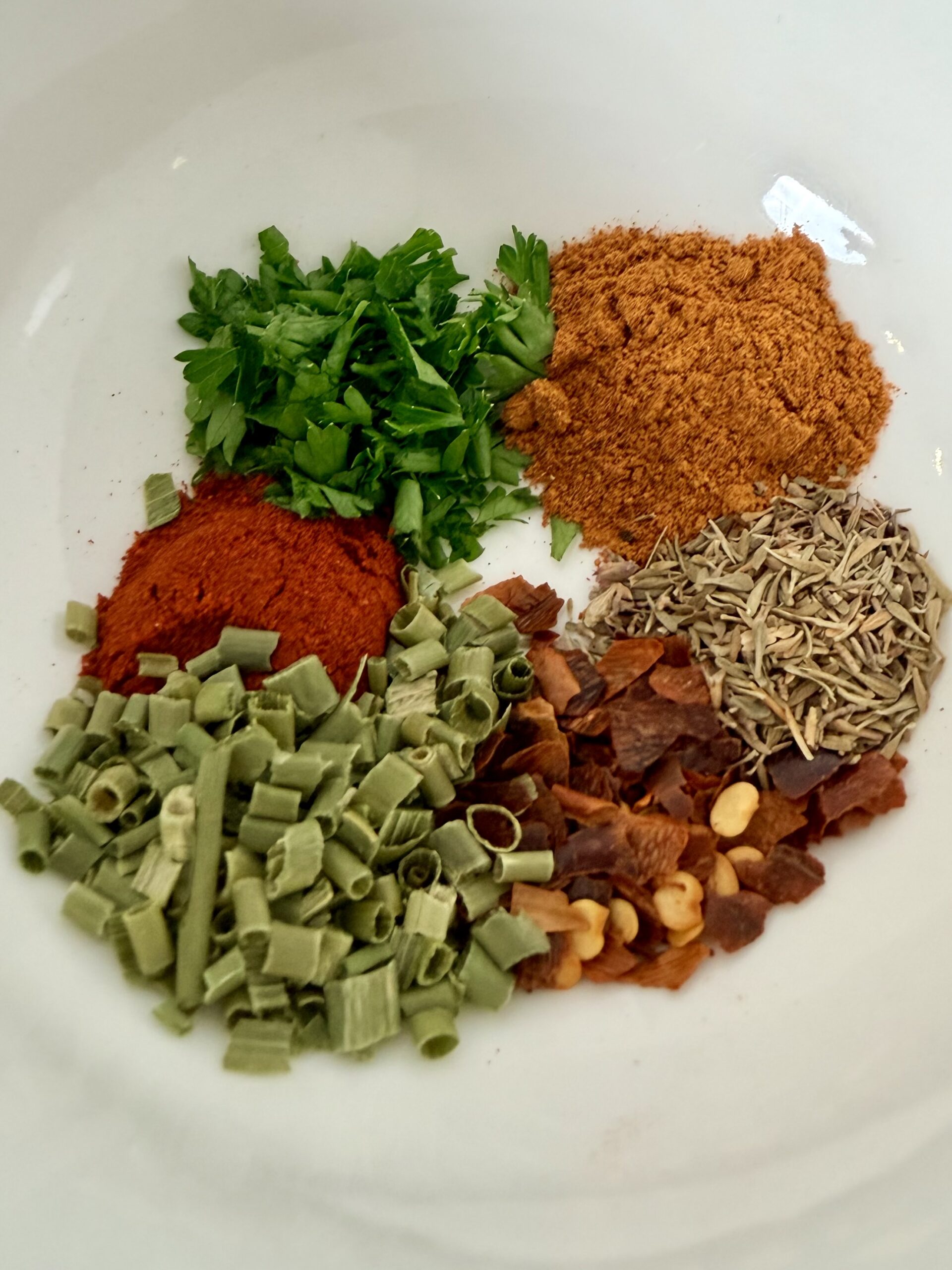 A white plate with six piles of different spices and herbs, including chopped parsley, paprika, curry powder, dried chives, dried thyme, and crushed red pepper flakes.