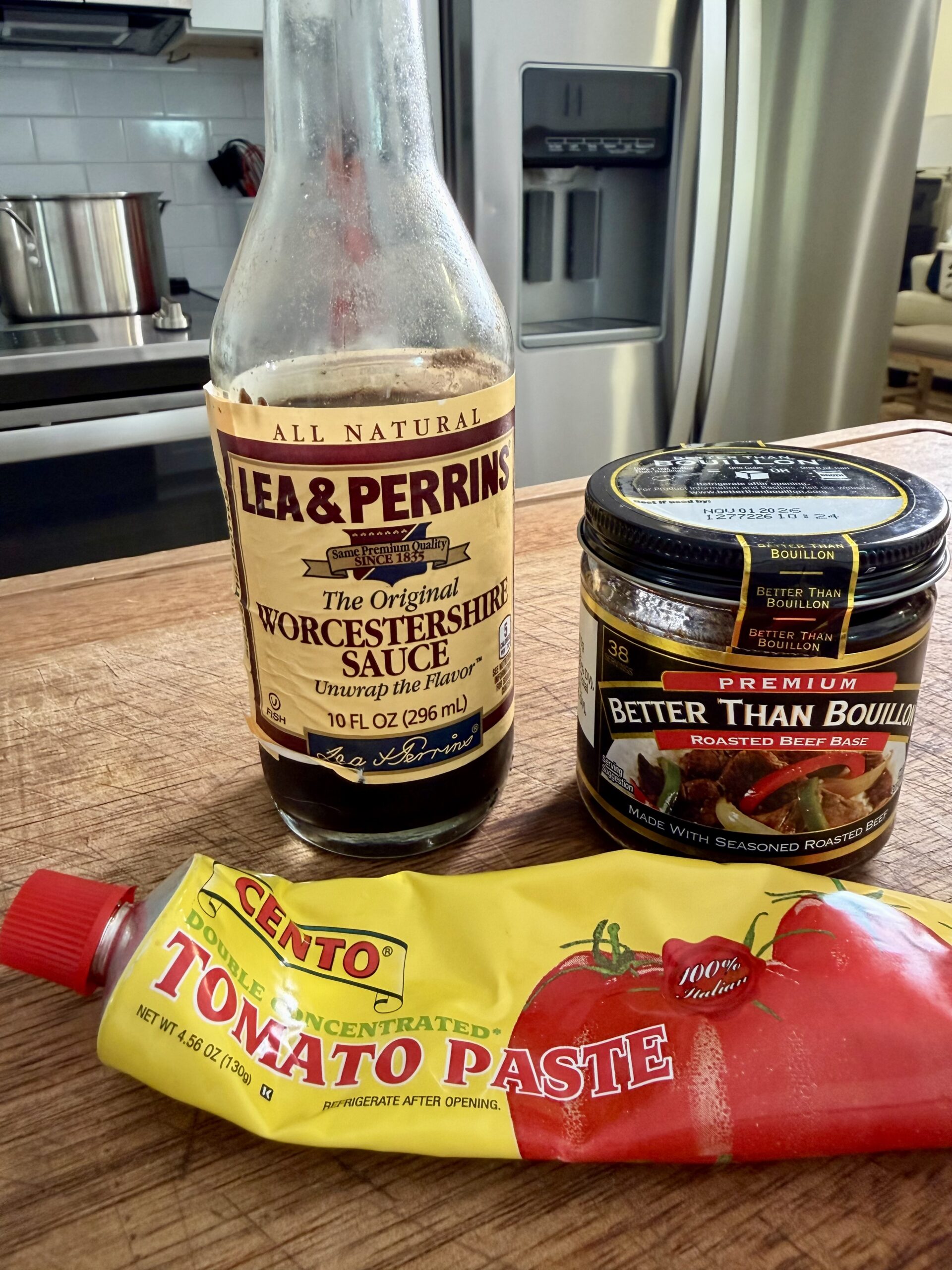 A bottle of Lea & Perrins Worcestershire Sauce, a jar of Better Than Bouillon Roasted Beef Base, and a tube of Cento Tomato Paste are arranged on a kitchen counter with pots and appliances in the background.