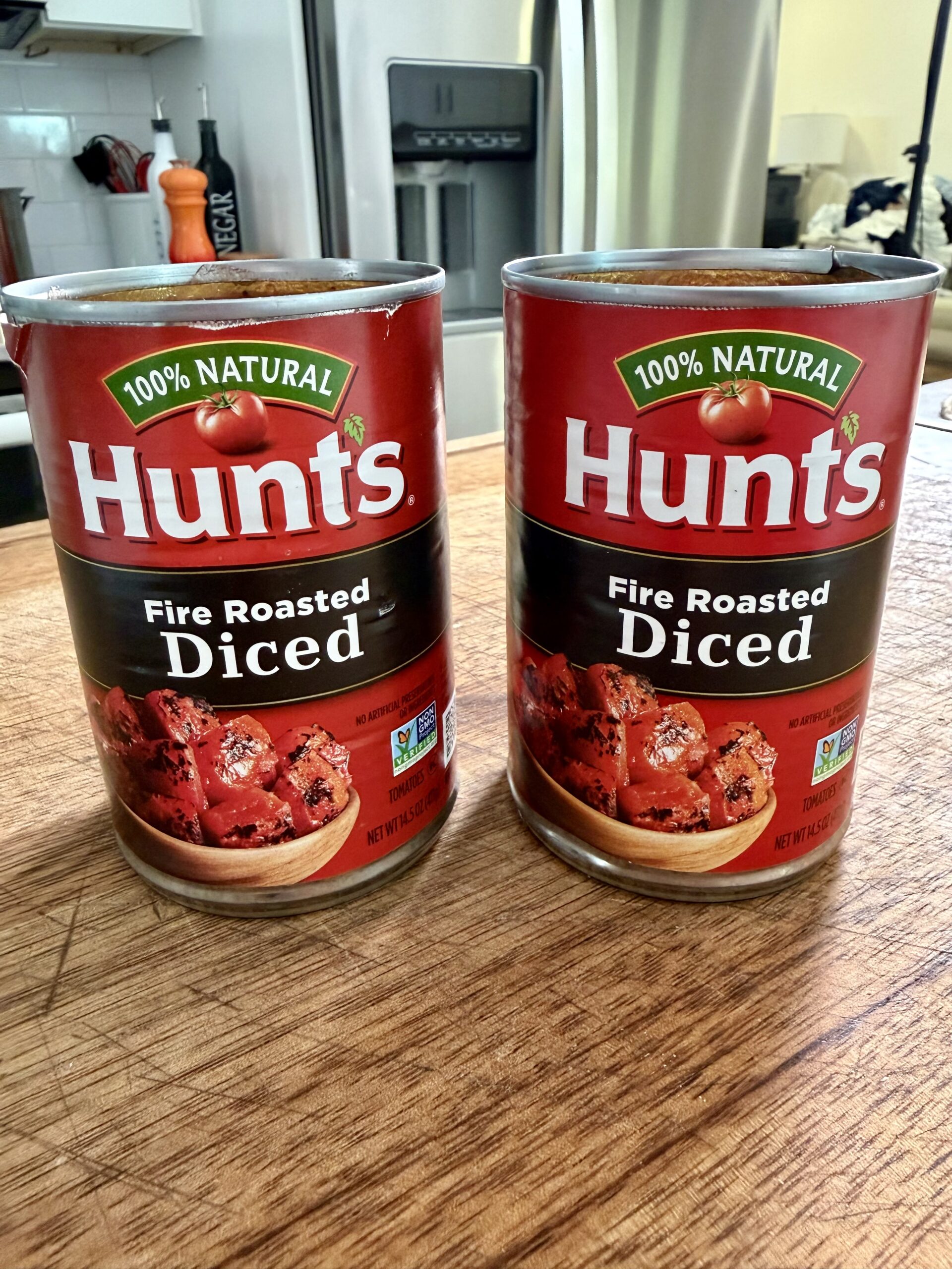 Two cans of Hunt’s Fire Roasted Diced Tomatoes sit on a wooden countertop in a kitchen with a refrigerator and various kitchen items in the background.