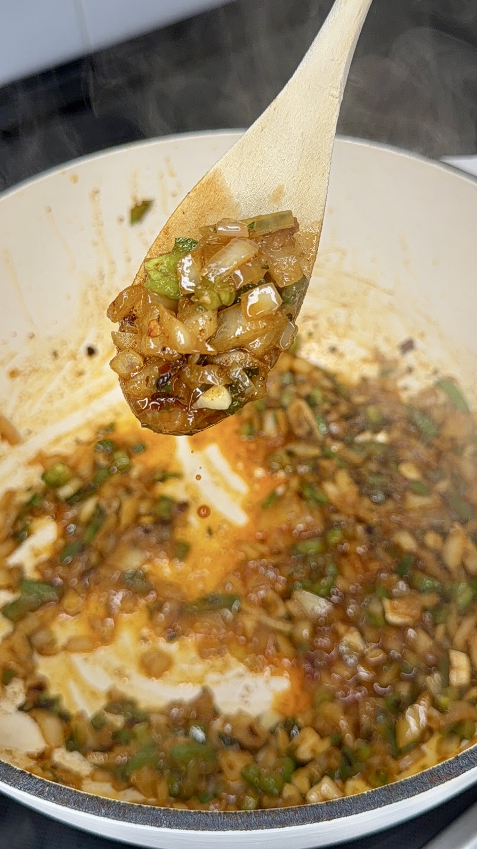 A wooden spoon holds a mix of sautéed onions, green peppers, and spices above a pot filled with the same sizzling, colorful vegetable mixture. Steam rises from the pot, suggesting the food is hot and freshly cooked.