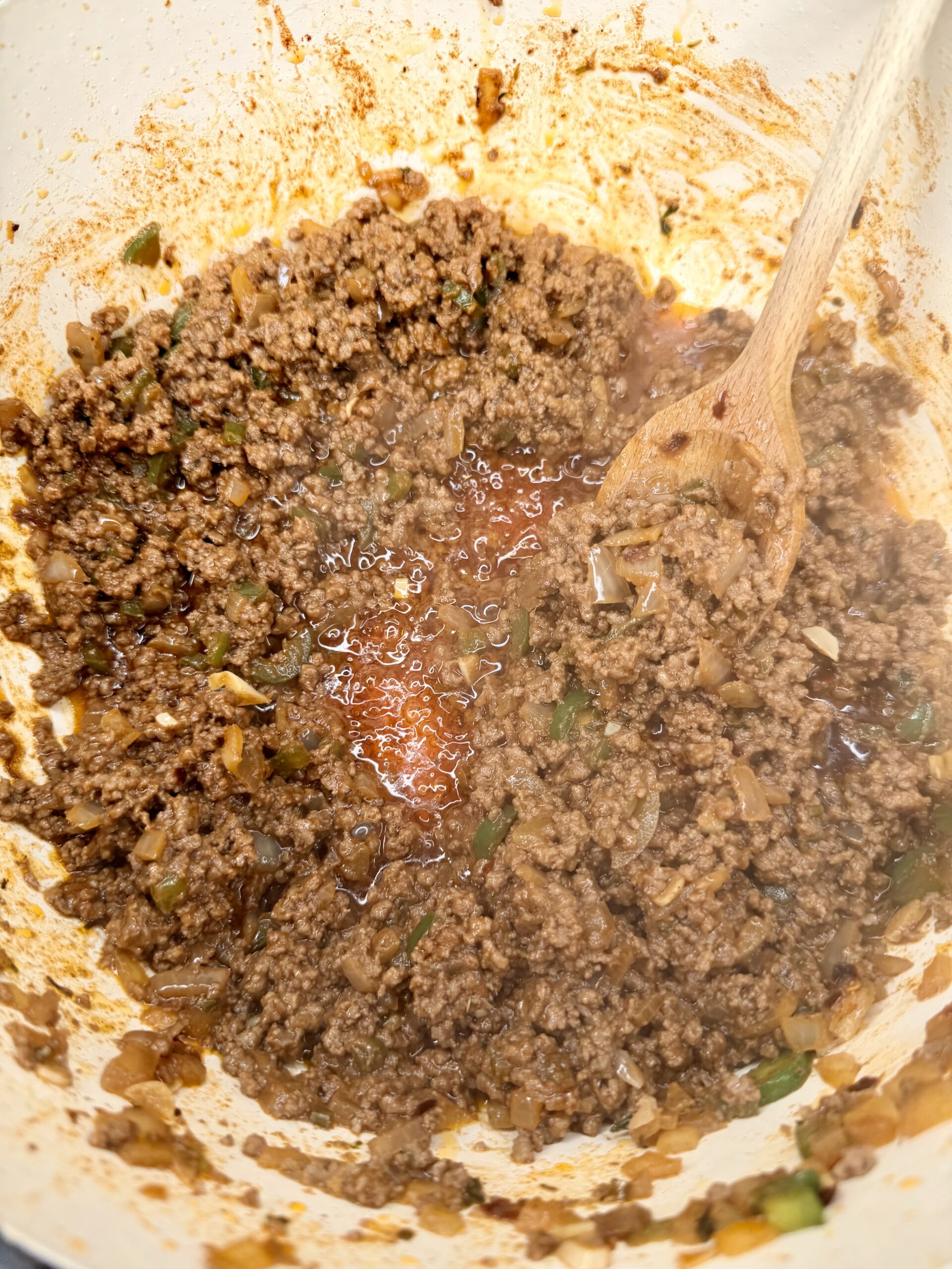 Ground beef, chopped onions, and green peppers being cooked and mixed in a large white pot with a wooden spoon; the mixture is browned and releasing juices.
