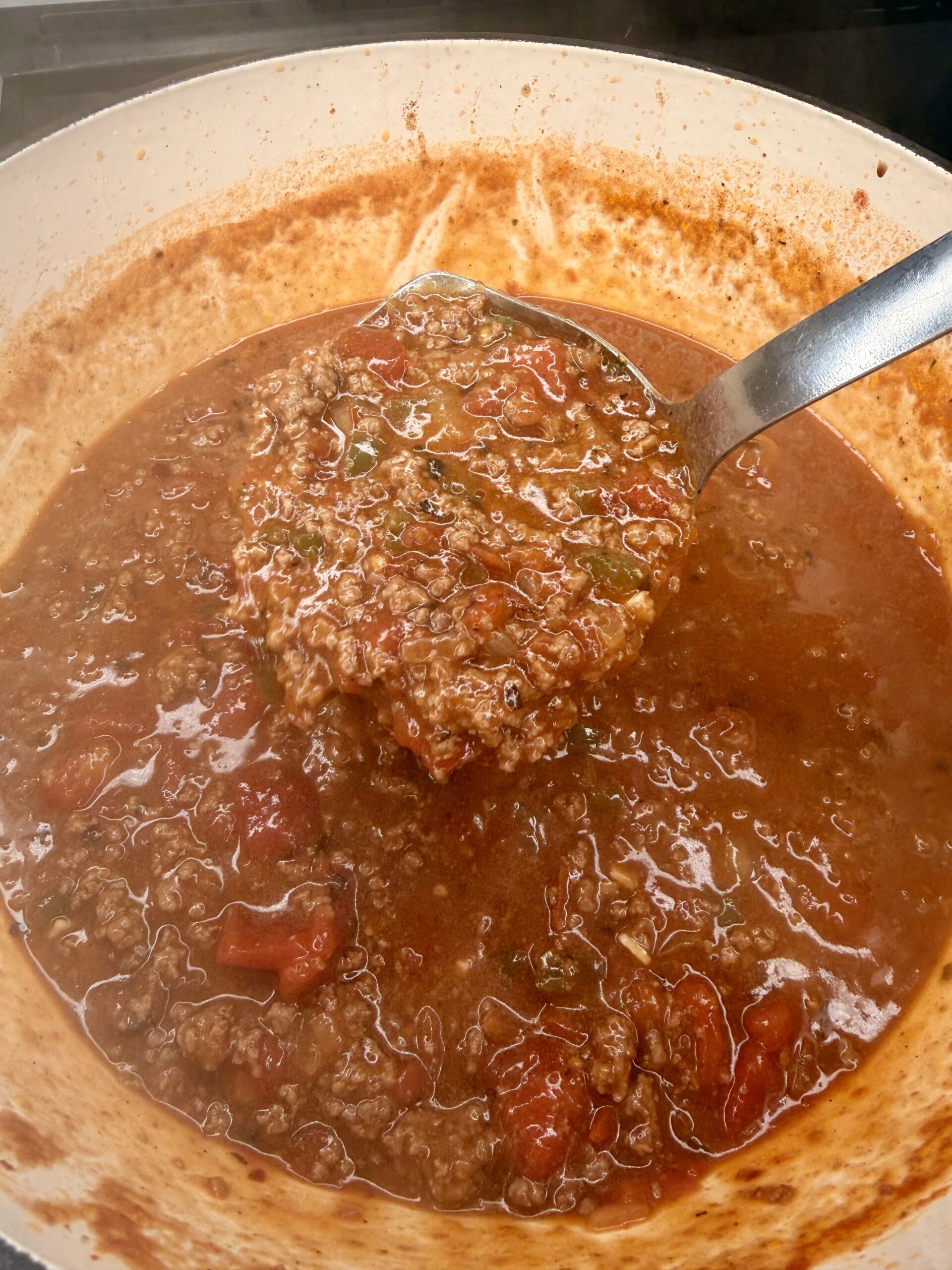 A close-up of a large pot of simmering meat sauce with ground beef, tomatoes, and herbs. A metal ladle is lifting a hearty scoop of the chunky, red sauce above the pot.