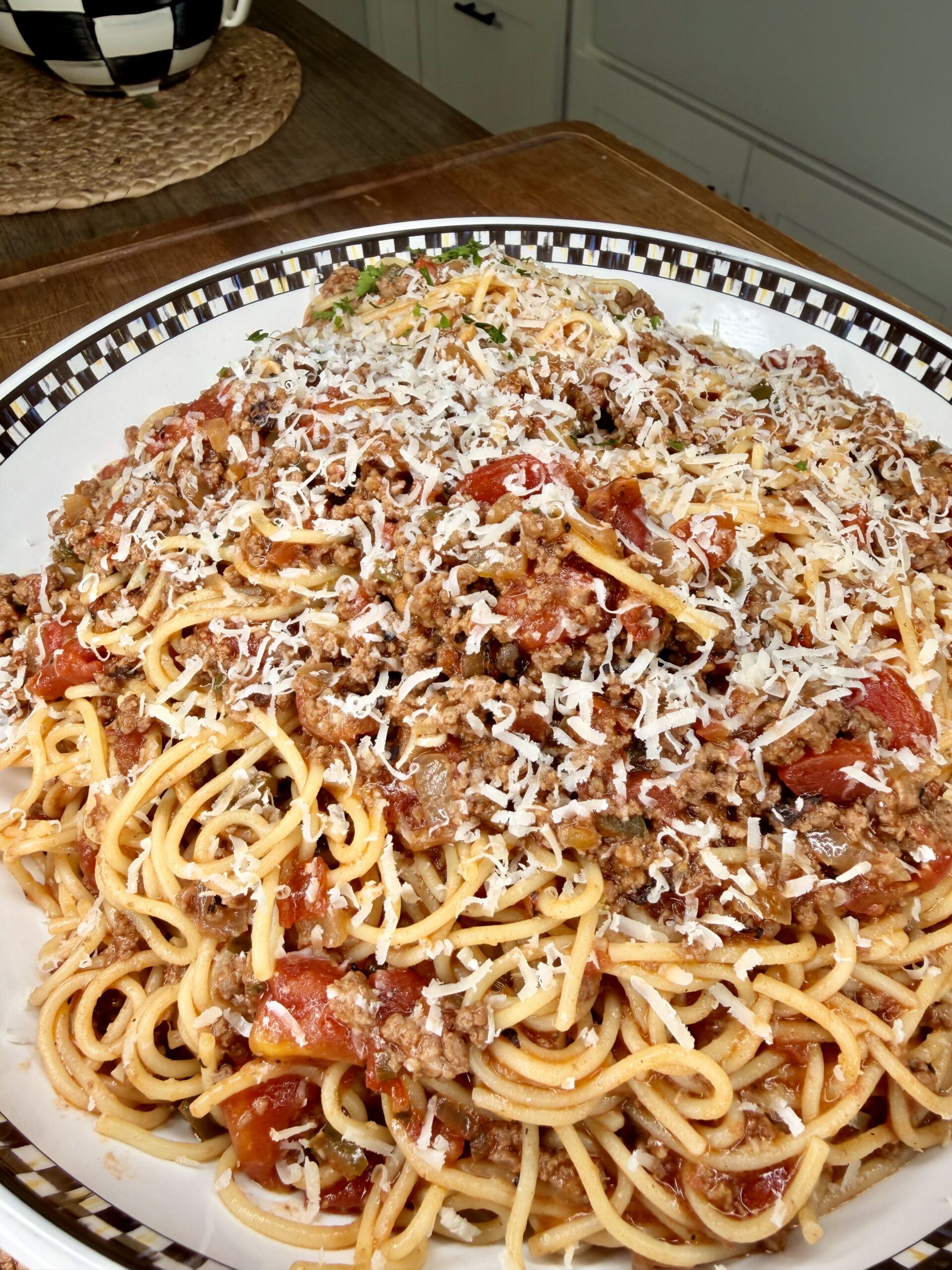 A large plate of spaghetti topped with meat sauce and grated cheese, served on a checkered-rim dish on a kitchen table.