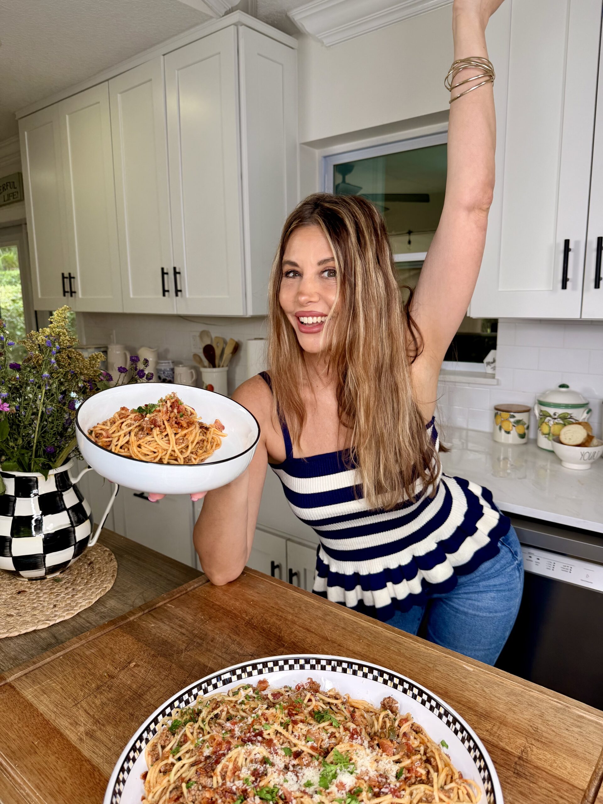 A smiling woman in a striped tank top holds up a bowl of pasta with one hand raised, standing in a bright kitchen with white cabinets. A plate of pasta is on the wooden counter in front of her.