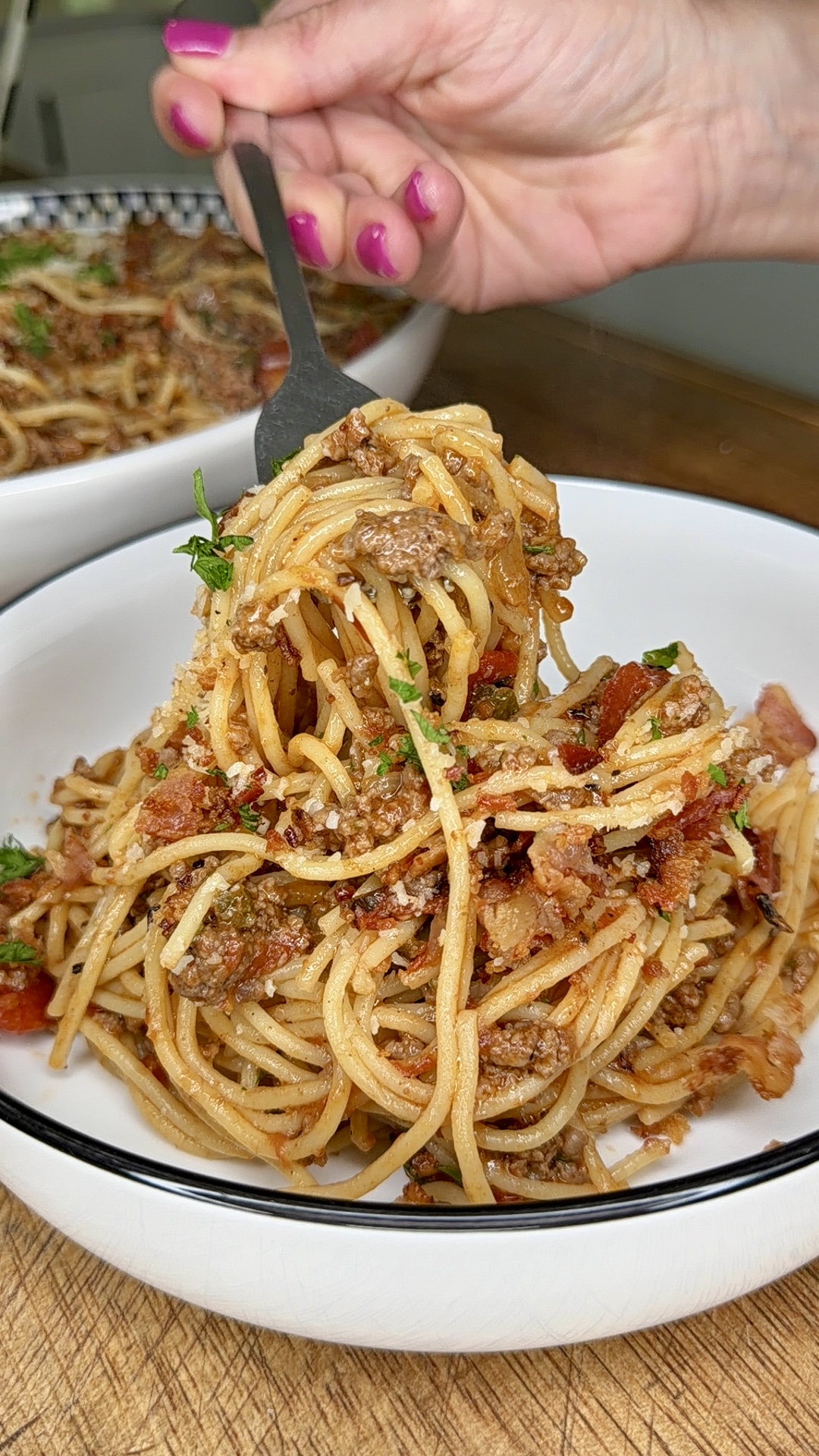 A hand with pink nails twirls a forkful of spaghetti with meat sauce and herbs in a white bowl set on a wooden surface. Another bowl of pasta is visible in the background.