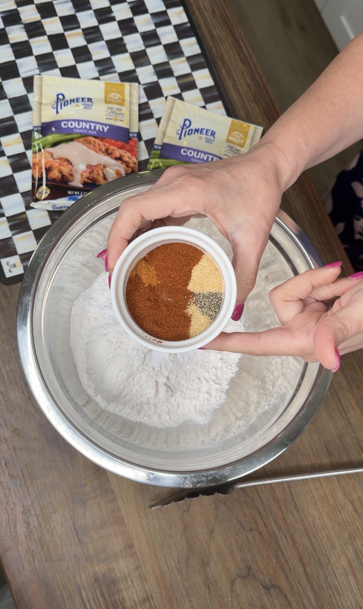 A person holds a small bowl of assorted spices over a large mixing bowl with flour. Two packets of Pioneer Country Gravy Mix and a checked cloth are on the table nearby.
