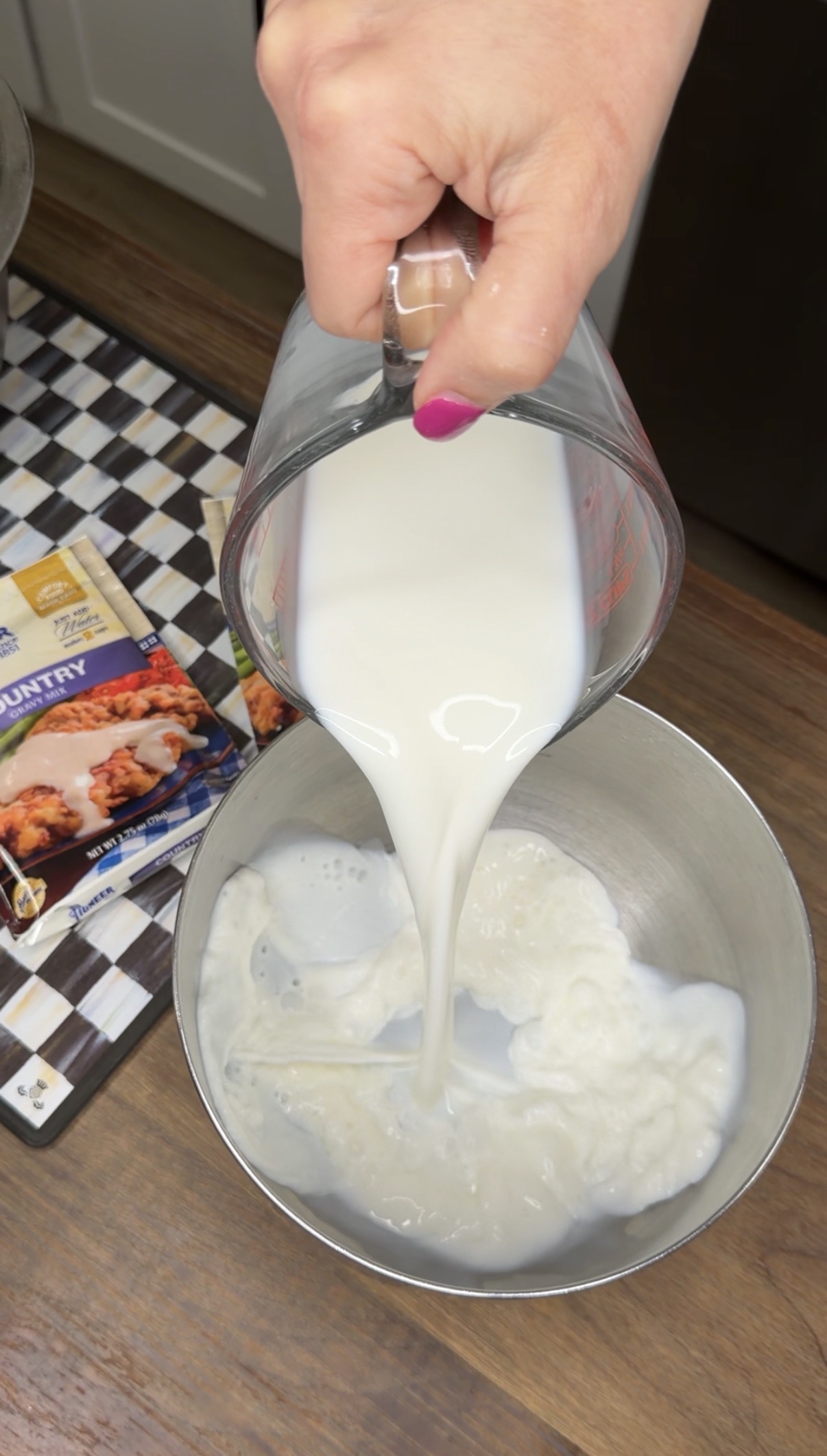 A hand with pink nail polish pours milk from a glass measuring cup into a metal mixing bowl. A boxed mix rests on a checkered tray nearby on a wooden surface.