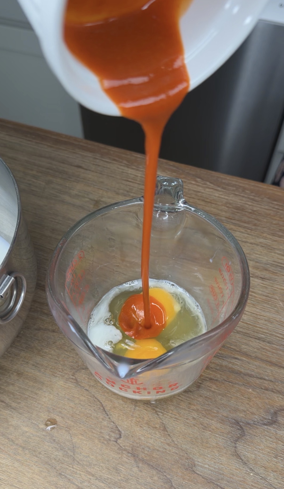 A stream of orange-red sauce is being poured into a glass measuring cup containing raw eggs, on a wooden countertop next to a metal pot.