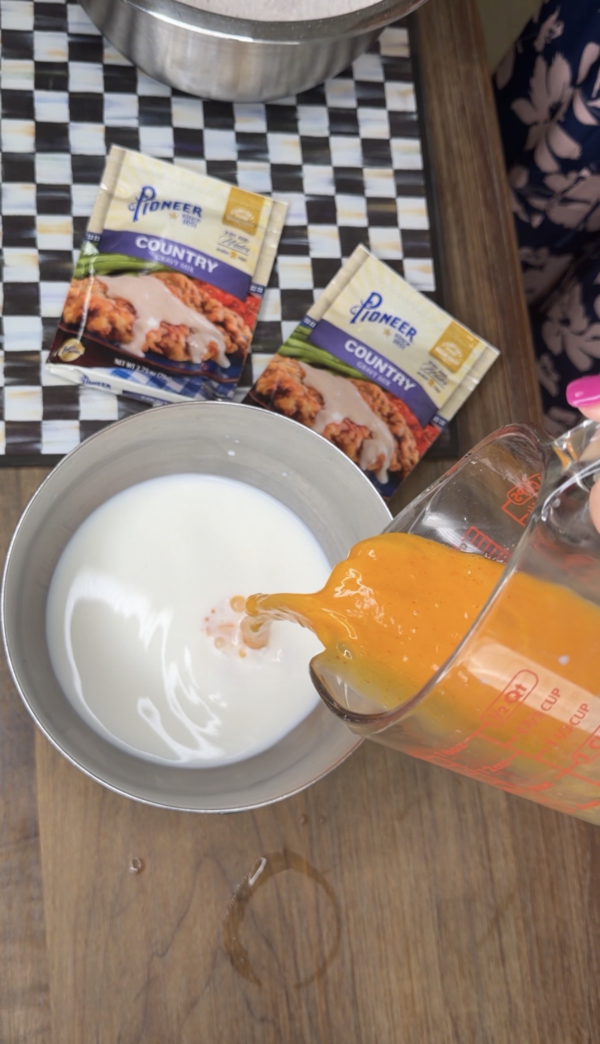 A hand pours an orange liquid from a measuring cup into a bowl of milk. Two packets of Pioneer Country Gravy Mix are on the table nearby.