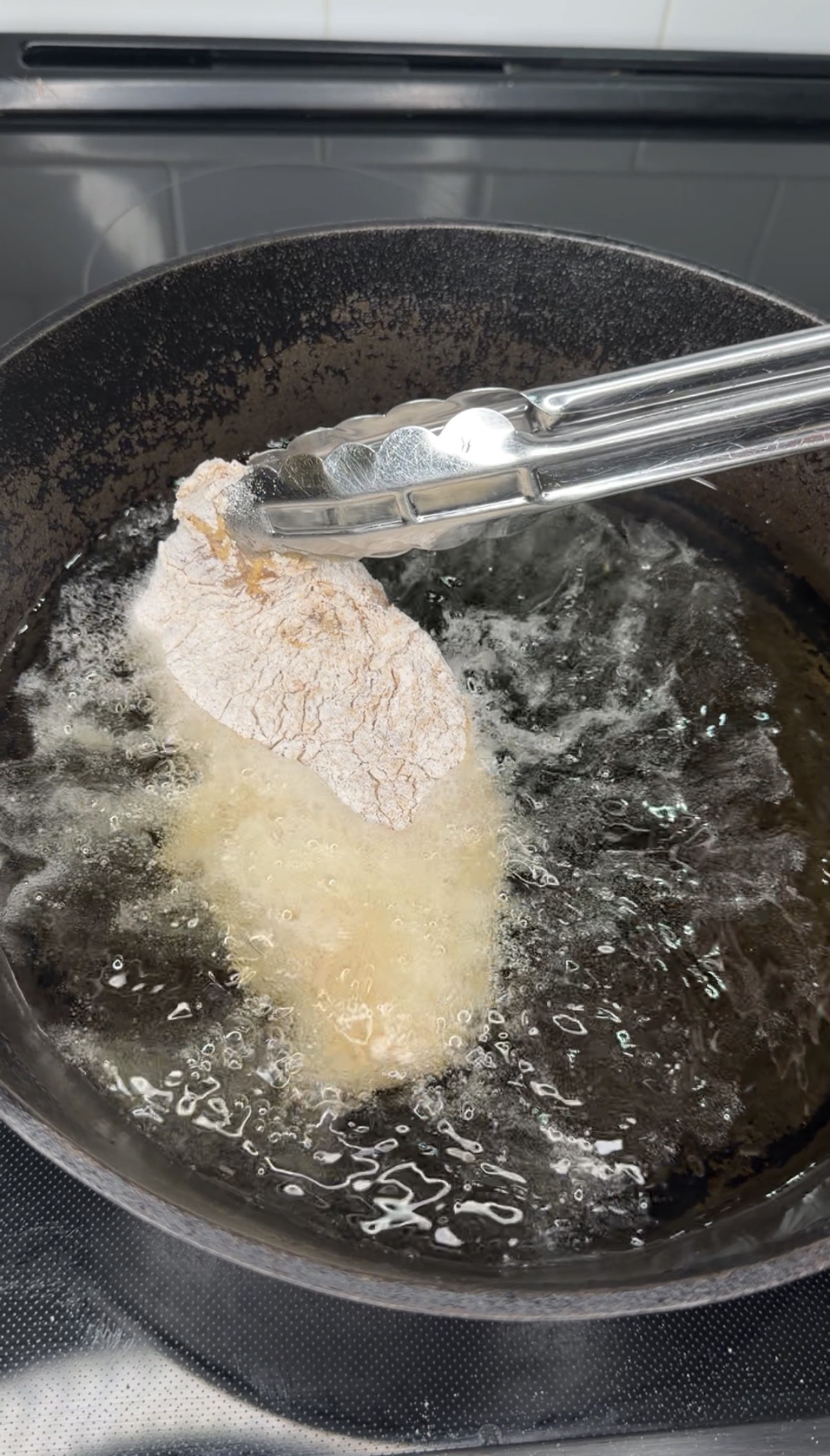 A metal tong holds a piece of breaded chicken as it is lowered into hot oil in a black frying pan, with bubbles forming around the chicken as it starts to fry.