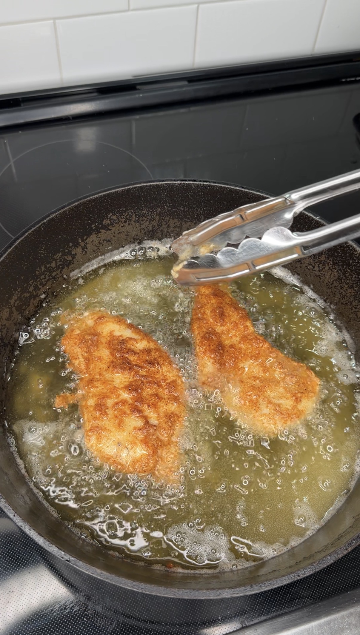 Two breaded chicken cutlets frying in hot oil in a black skillet, with a pair of metal tongs holding one piece, on a stovetop with white tile backsplash.