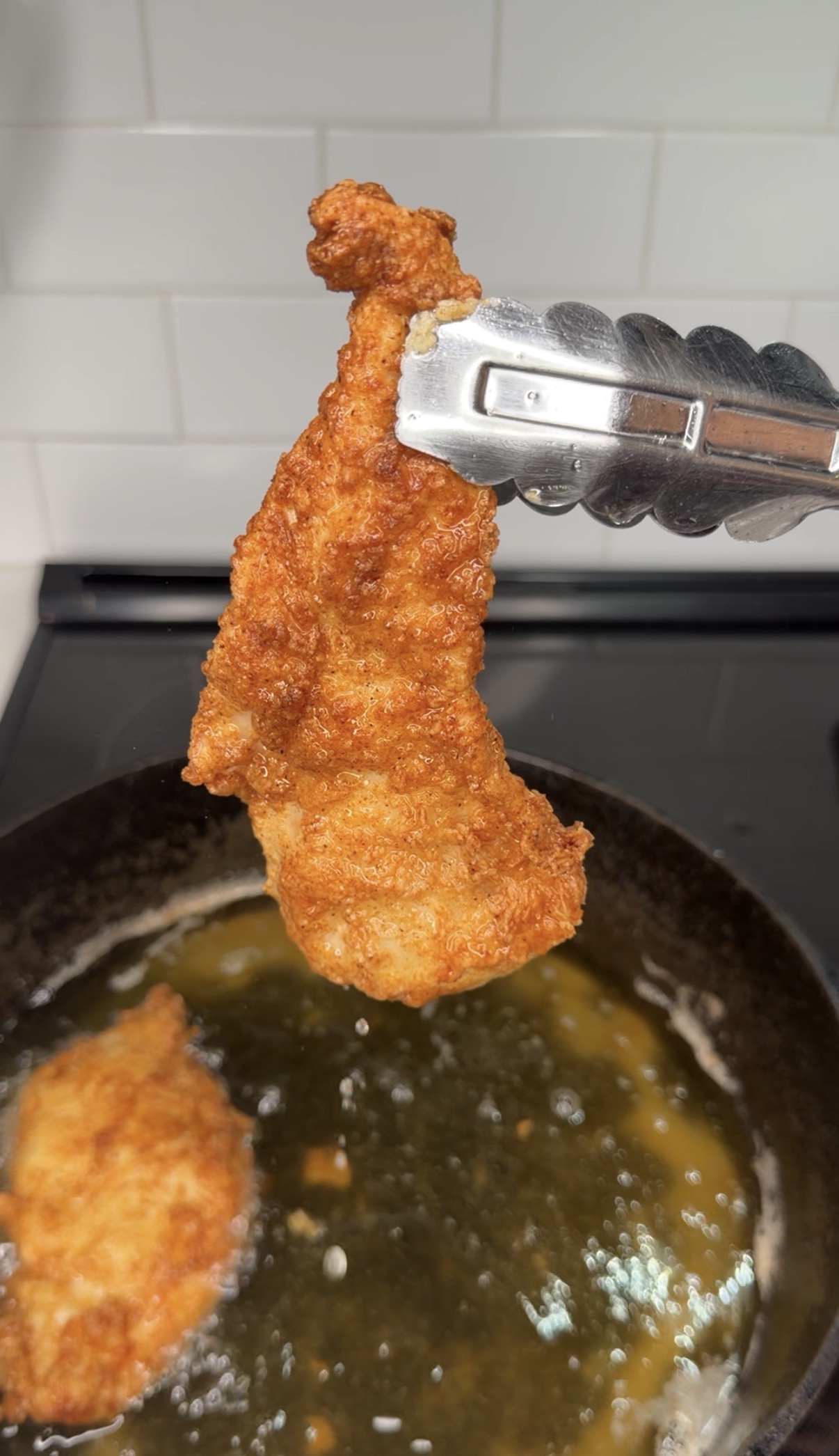 A piece of golden brown fried chicken is held above a frying pan with metal tongs. Another piece of chicken is frying in bubbling oil below. The kitchen background includes a stovetop and white tile backsplash.