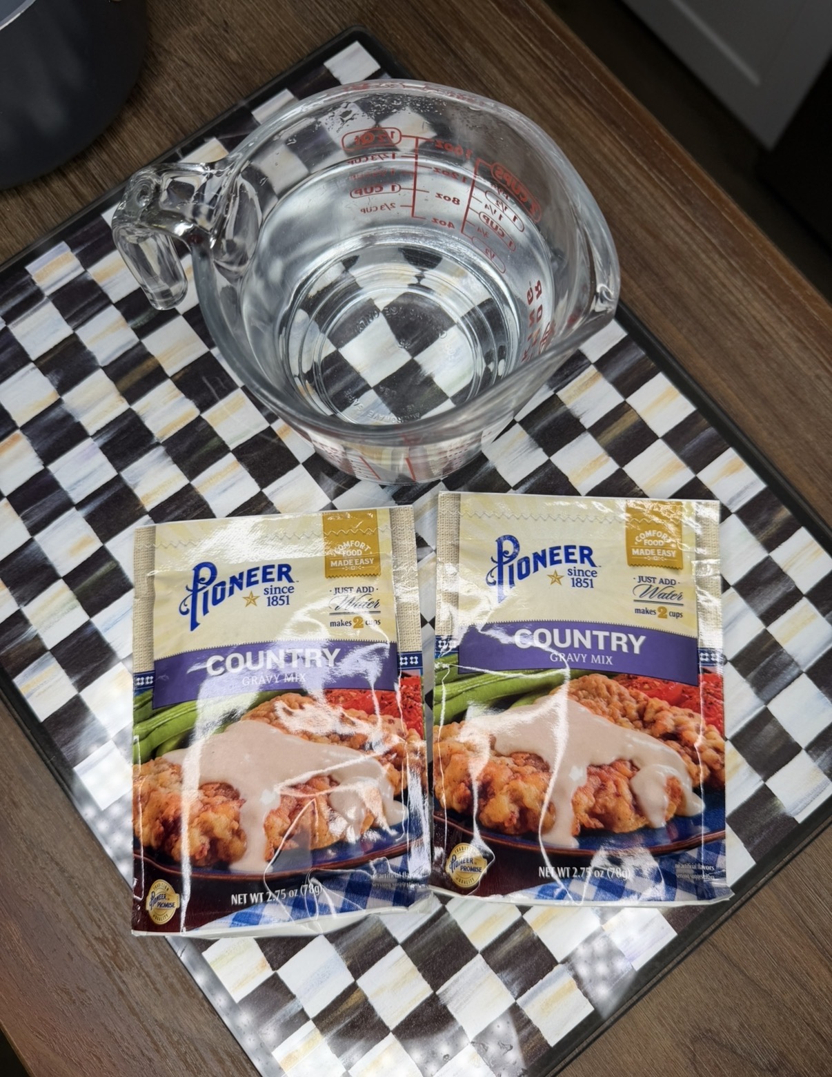 A glass measuring cup filled with water sits on a checkered surface next to two packets of Pioneer Brand Country Gravy Mix.