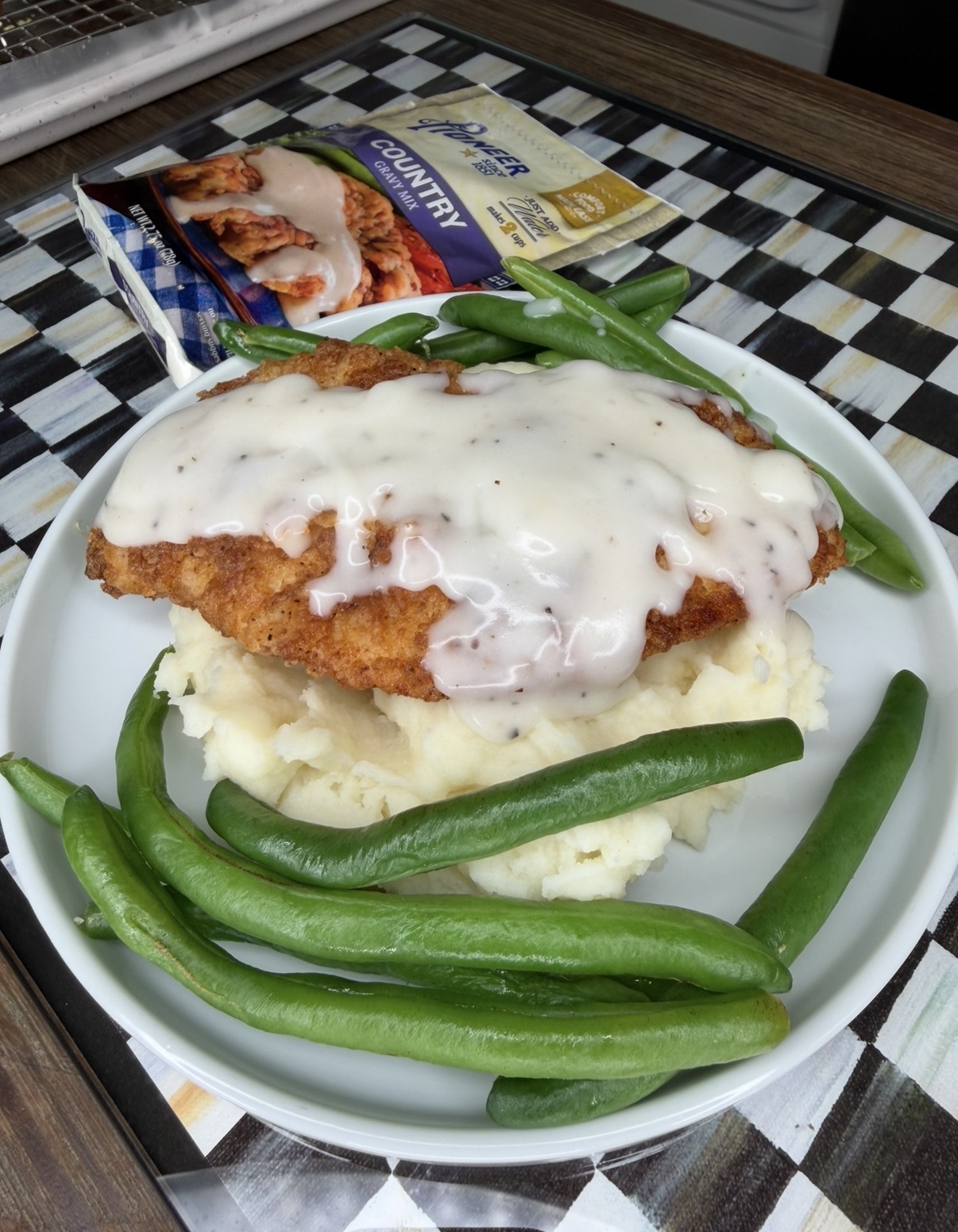 A plate of mashed potatoes topped with breaded chicken and white gravy, surrounded by green beans. The meal is next to a box labeled Banquet Country Fried Chicken Meal.