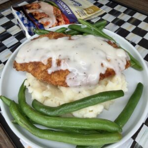 A plate with mashed potatoes topped with breaded chicken and white gravy, surrounded by green beans. In the background, there is a box of frozen country fried chicken meal.