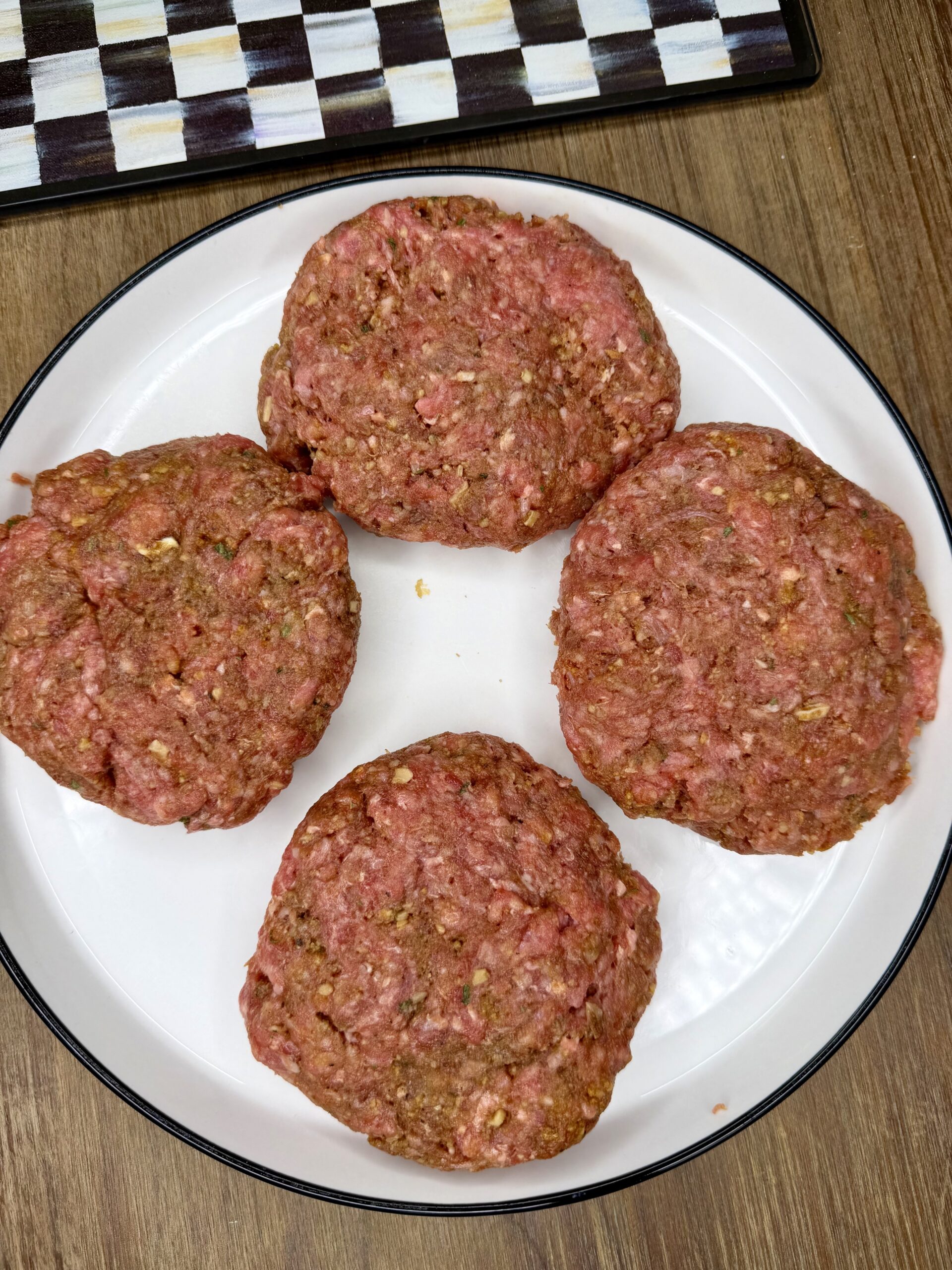 Four raw ground beef burger patties arranged in a circle on a white plate, placed on a wooden table with part of a checkered-patterned tray visible in the background.