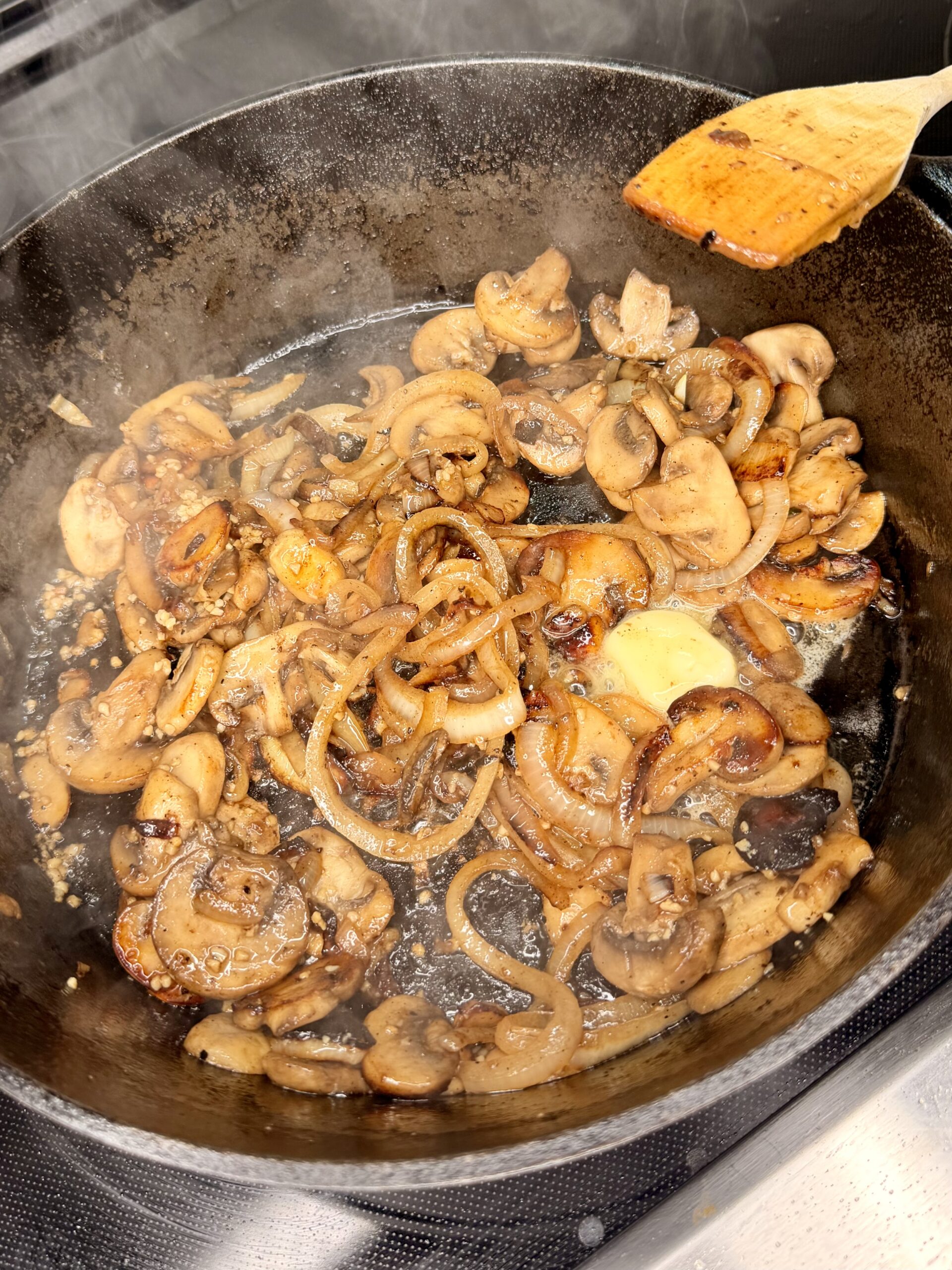 Sliced mushrooms and onions sautéing in a cast iron skillet, with a pat of butter melting in the center and a wooden spatula resting on the side. Steam rises from the hot pan.