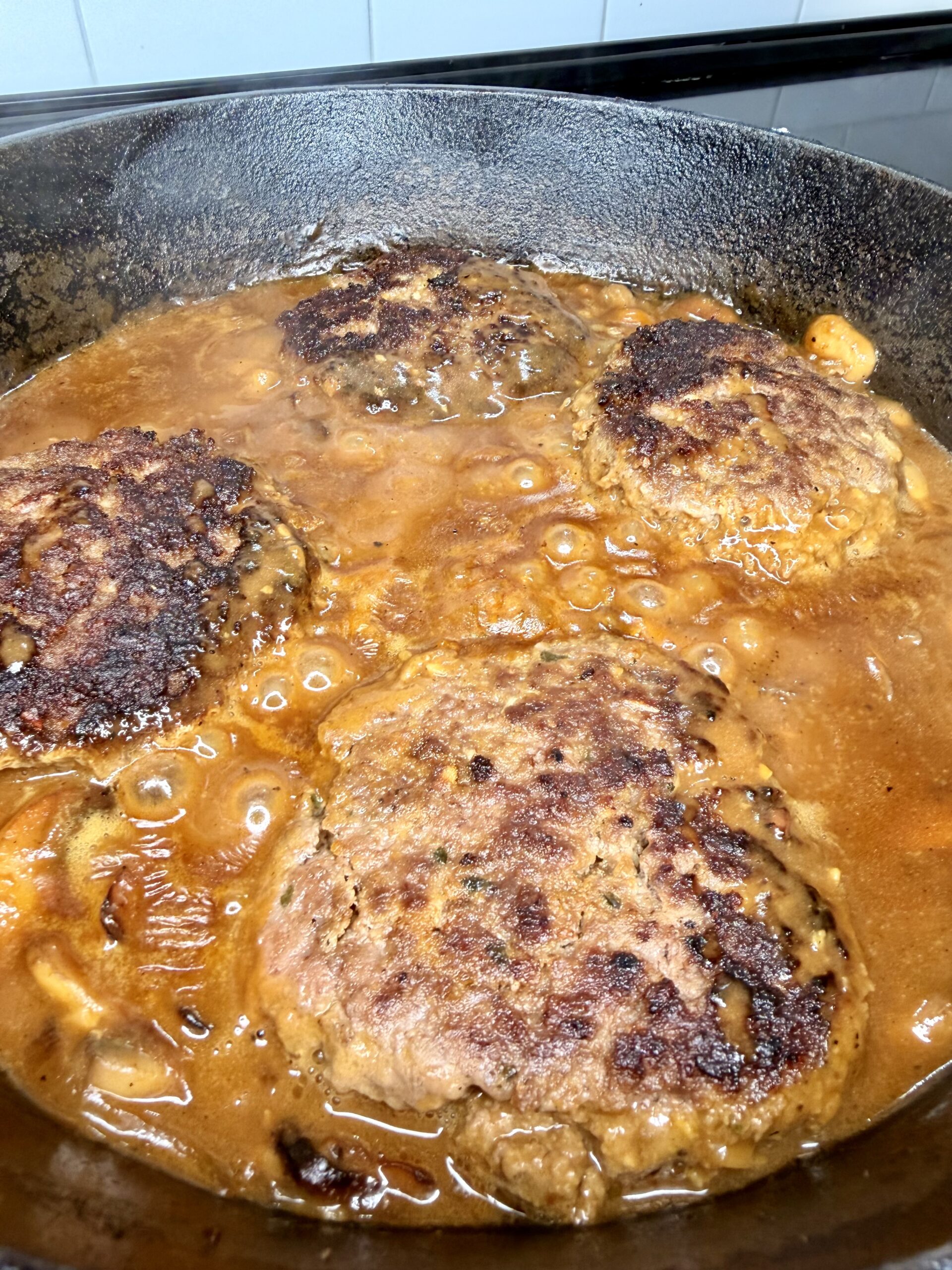 Four browned hamburger patties simmer in a thick, brown onion gravy inside a black cast iron skillet on a stovetop.