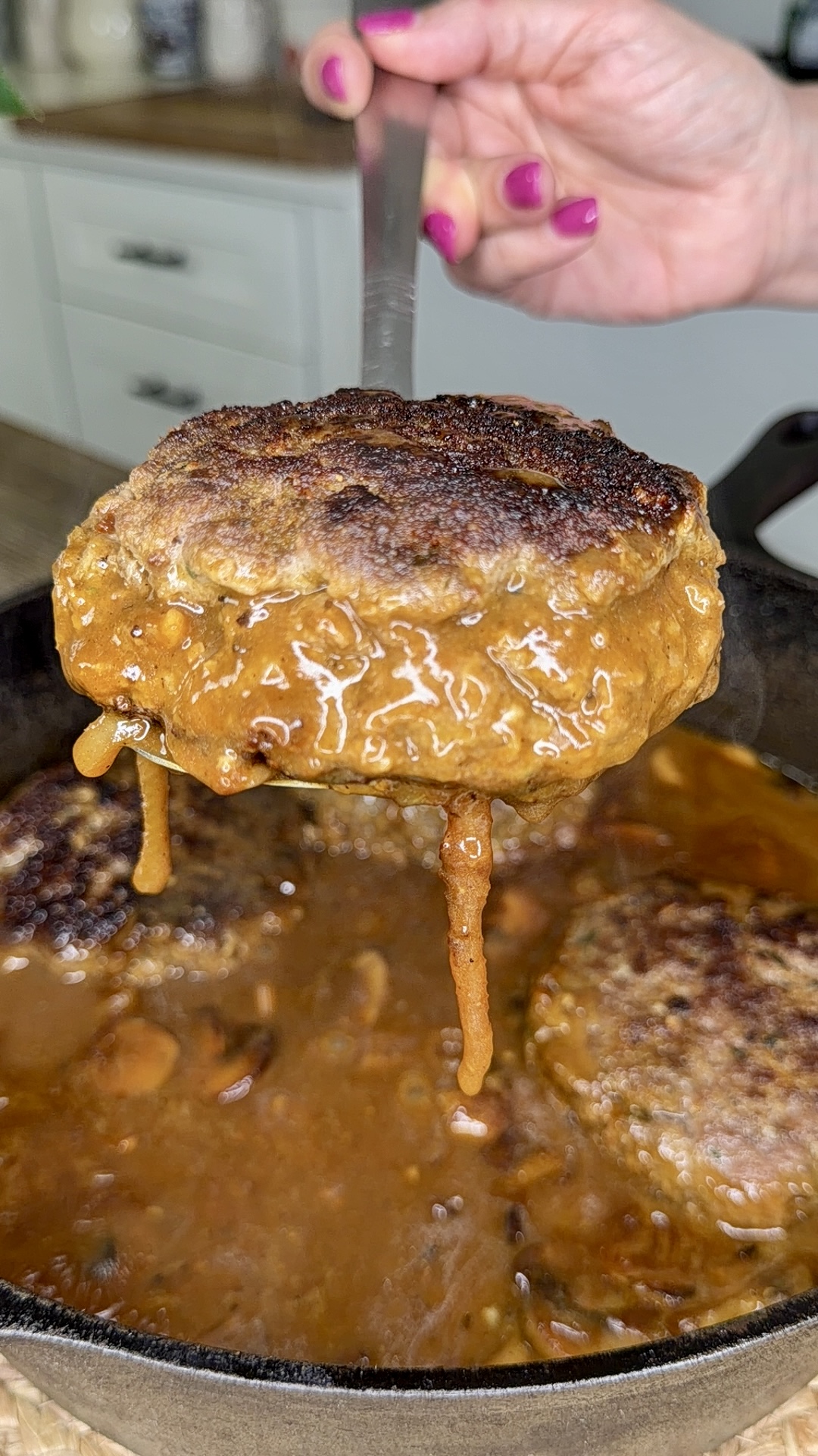 A hand with pink nail polish holds a spatula lifting a juicy, browned Salisbury steak covered in rich brown gravy from a cast iron skillet, with more steak and gravy visible inside the pan.