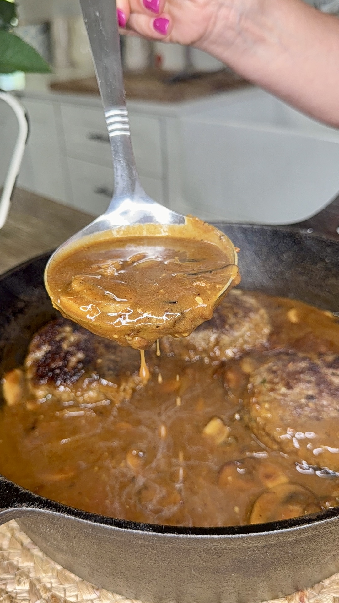A hand with pink nails holds a ladle of brown gravy over a cast iron skillet filled with meat patties and rich brown sauce, with steam rising from the pan in a kitchen setting.