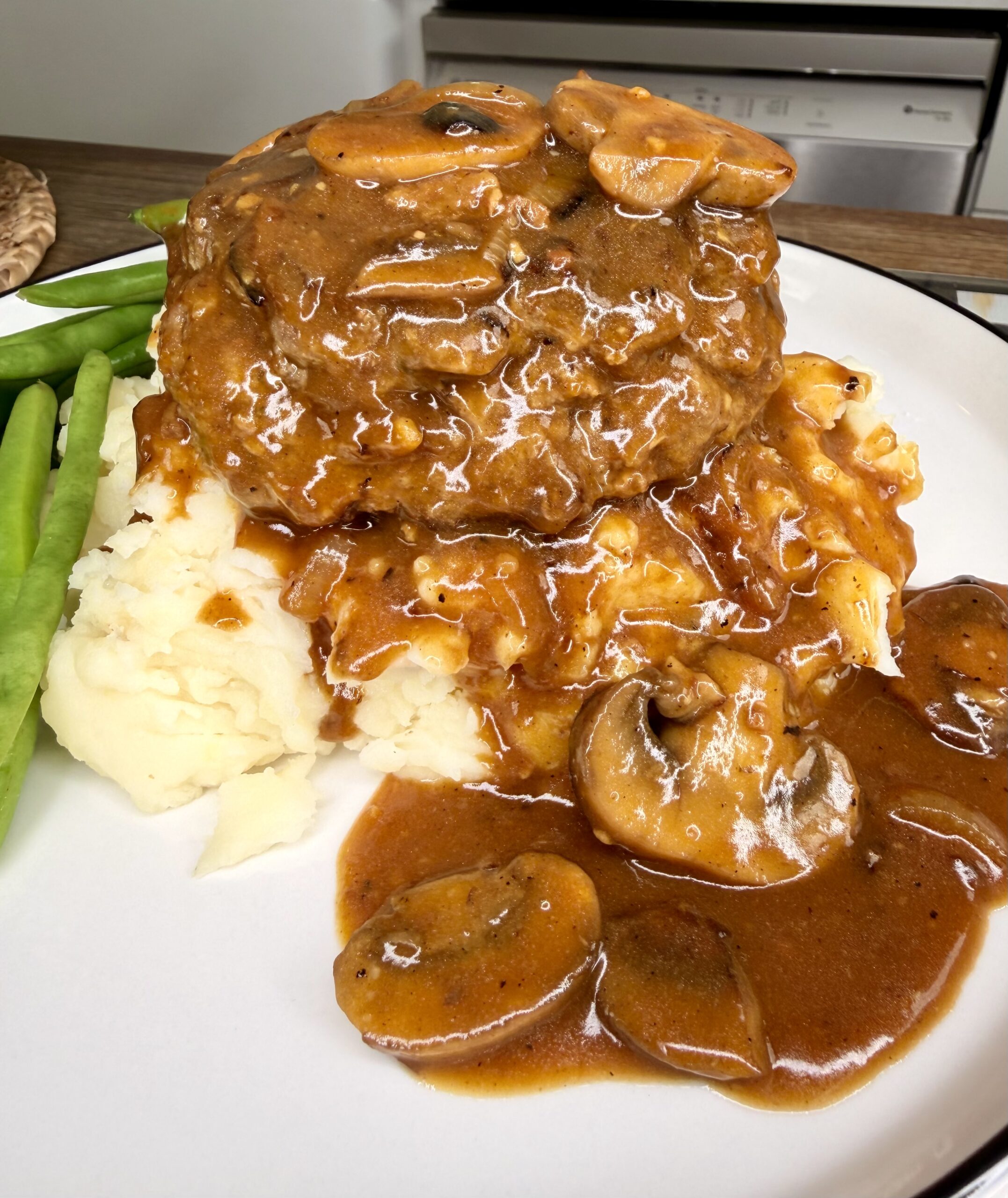 A Salisbury steak with mushrooms and brown gravy sits atop a mound of mashed potatoes, with extra gravy and mushrooms spilling onto the plate. Green beans are visible to the side.