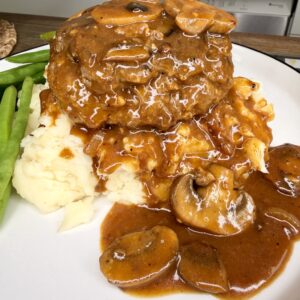 A serving of Salisbury steak with brown mushroom gravy sits on a heap of mashed potatoes, with gravy and mushrooms spilling onto the plate. Steamed green beans are on the side.