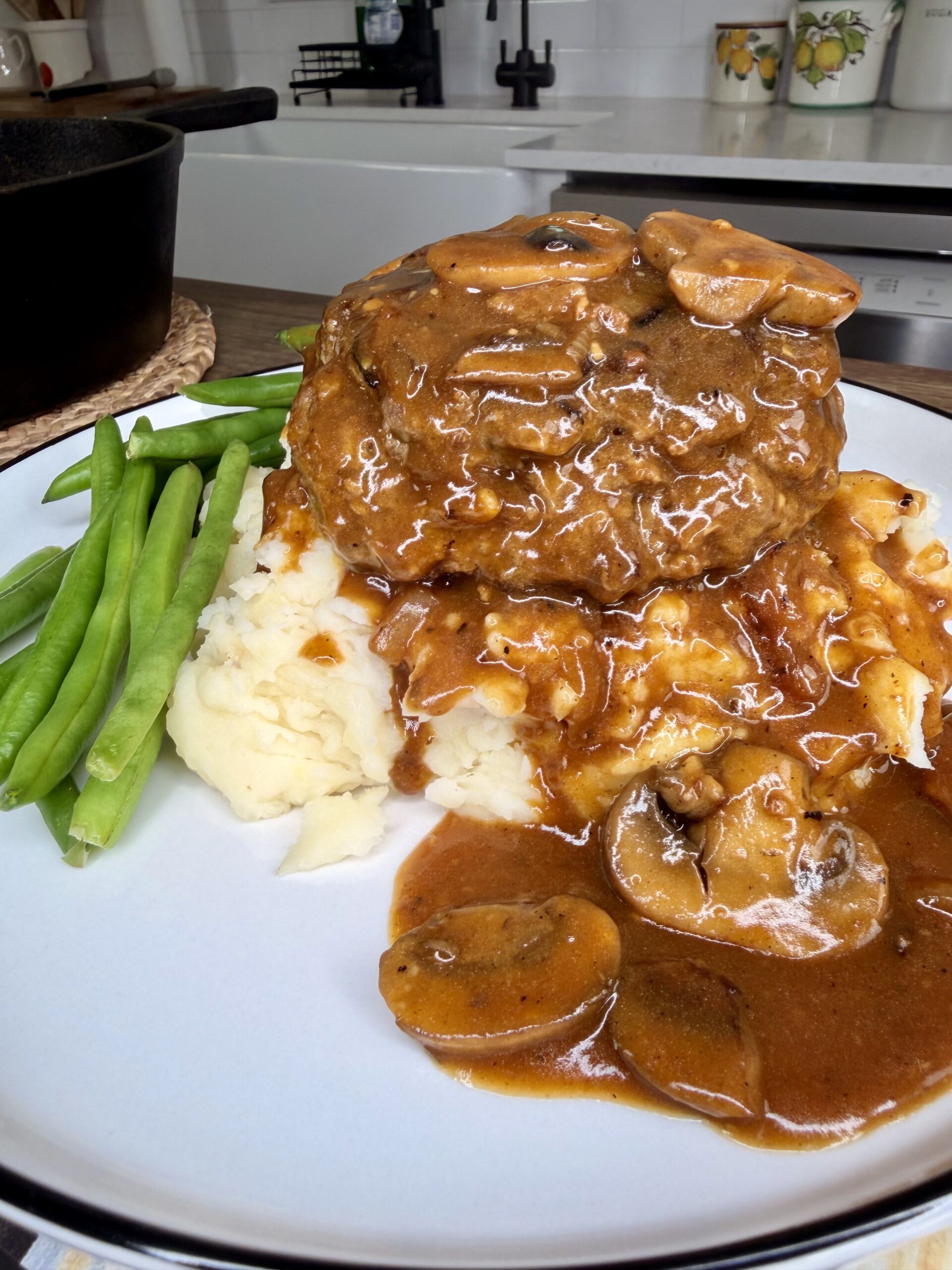 A plate of Salisbury steak topped with mushroom gravy, served over mashed potatoes with a side of green beans. The dish is in focus on a kitchen counter with a stove and sink in the background.