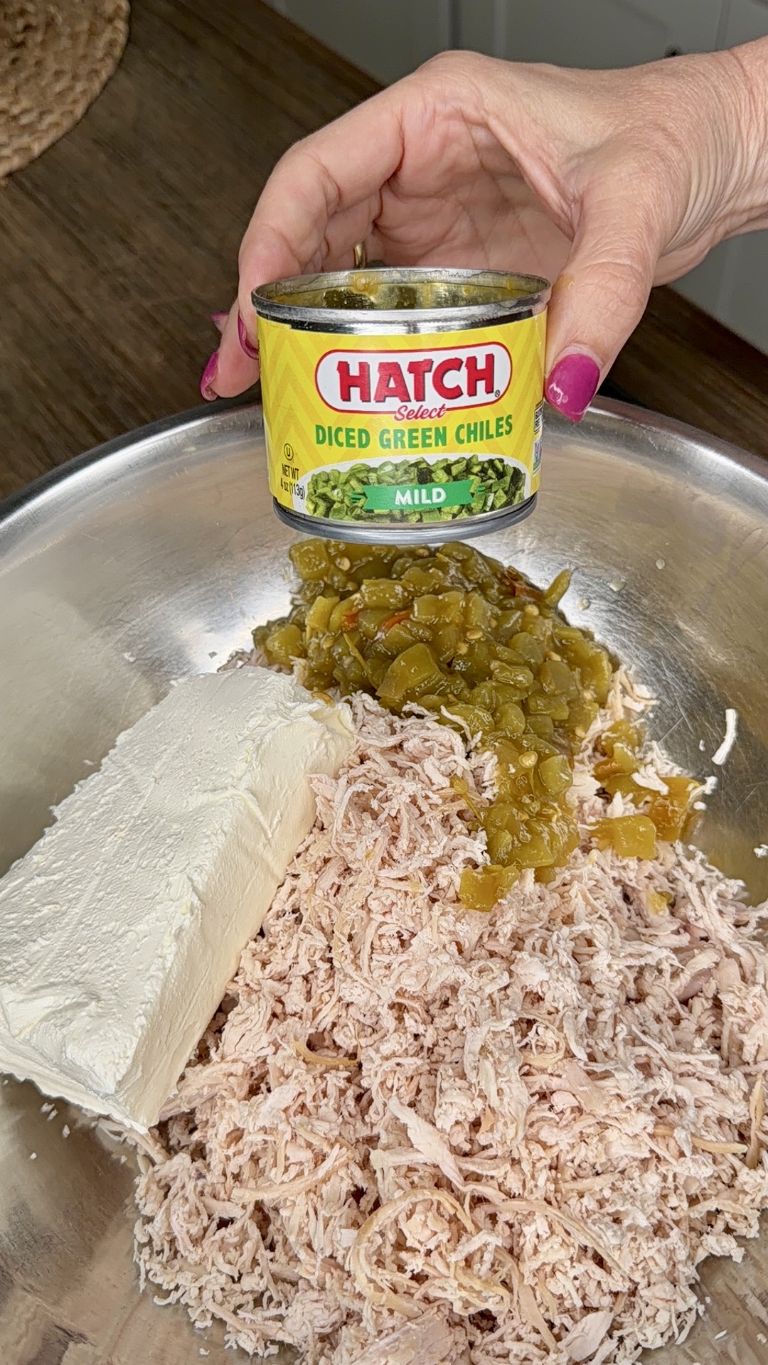 A hand holds a can of Hatch diced green chiles over a mixing bowl containing shredded chicken, cream cheese, and green chiles.
