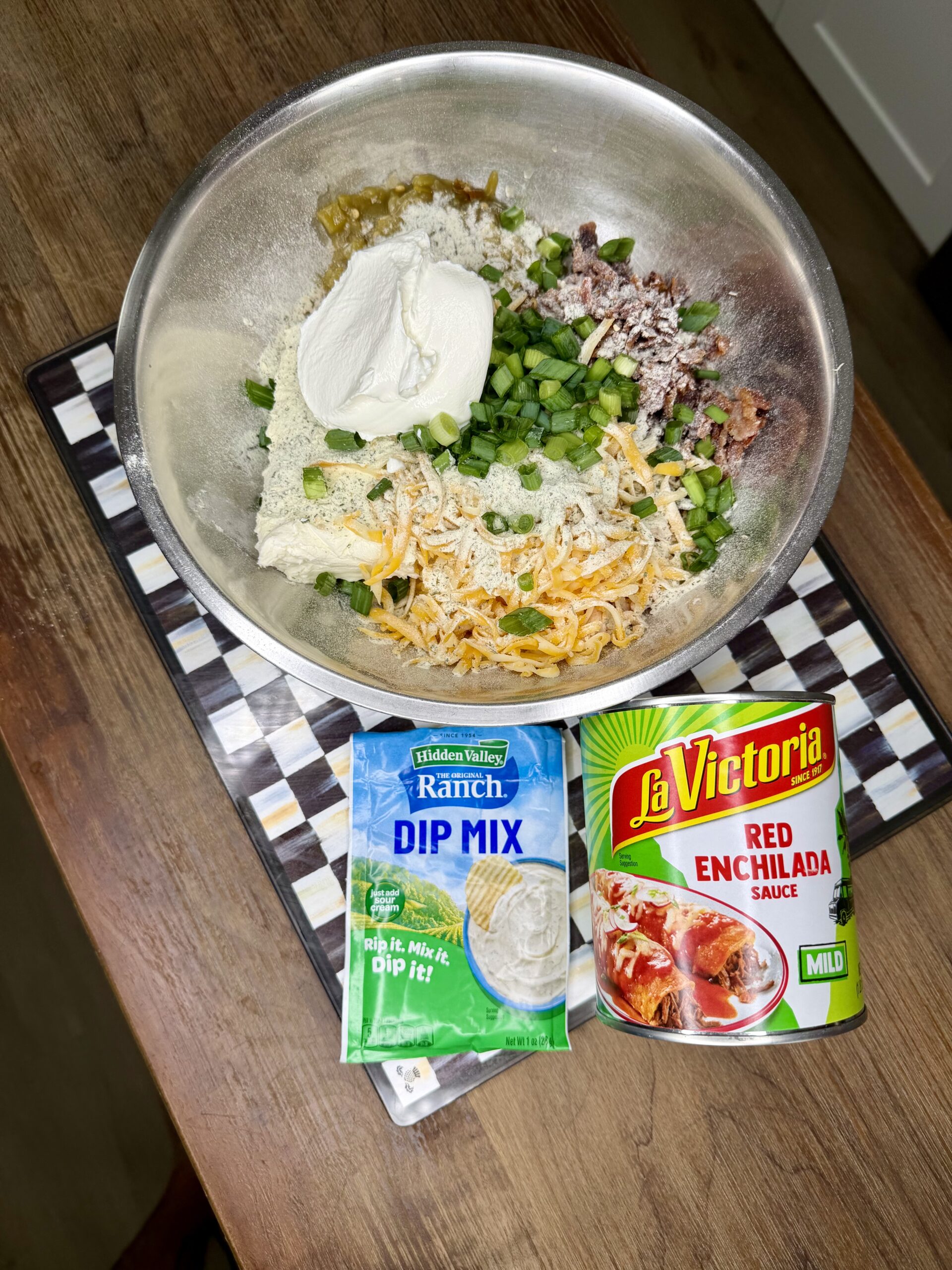 A bowl on a patterned tray holds dip ingredients: shredded cheese, cream cheese, sour cream, green onions, and chopped meat. In front are packets of Hidden Valley ranch dip mix and La Victoria red enchilada sauce.