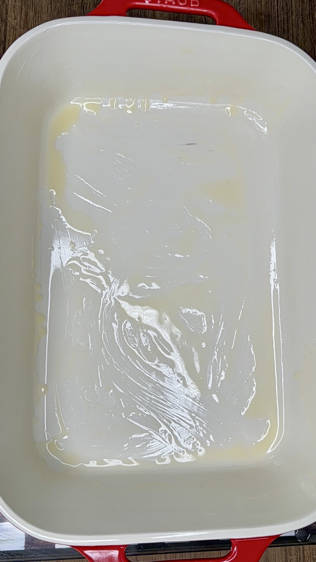A large white rectangular baking dish with red handles is coated with a thin layer of melted butter, visible as streaks on the surface. The dish sits on a wooden countertop.