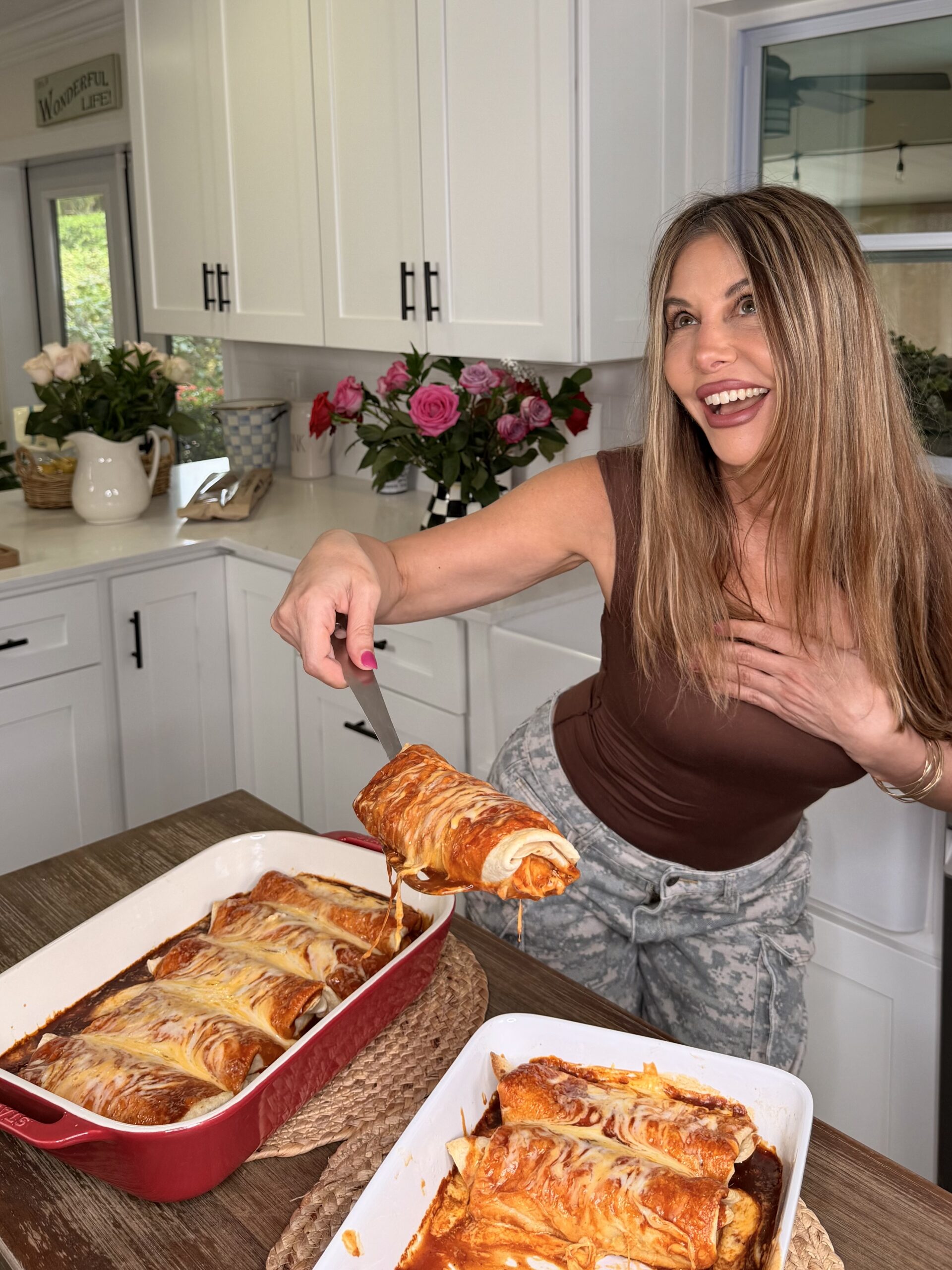 A smiling woman in a brown top serves Creamy Crack Chicken Enchiladas from a baking dish in a bright kitchen decorated with fresh flowers.