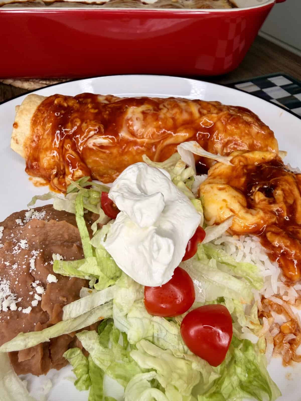 A plate with a cheese enchilada topped with red sauce, refried beans sprinkled with cheese, and a salad of shredded lettuce, cherry tomatoes, white rice, and a dollop of sour cream.