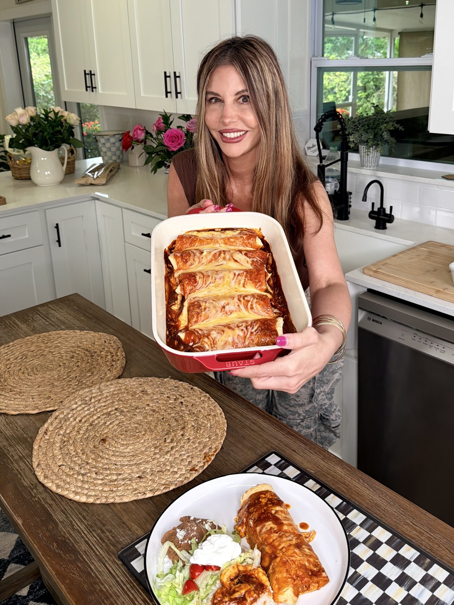 A woman in a bright kitchen smiles as she holds a baking dish of enchiladas. In the foreground, a plate with enchiladas, rice, beans, salad, and sour cream sits on a placemat on the counter.